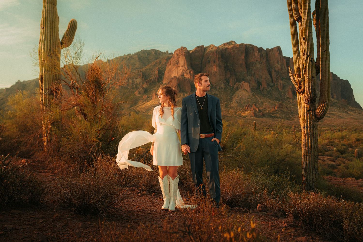 Couple in wedding or elopement attire standing among saguaro cacti with Superstition Mountains at golden hour in Arizona desert.
