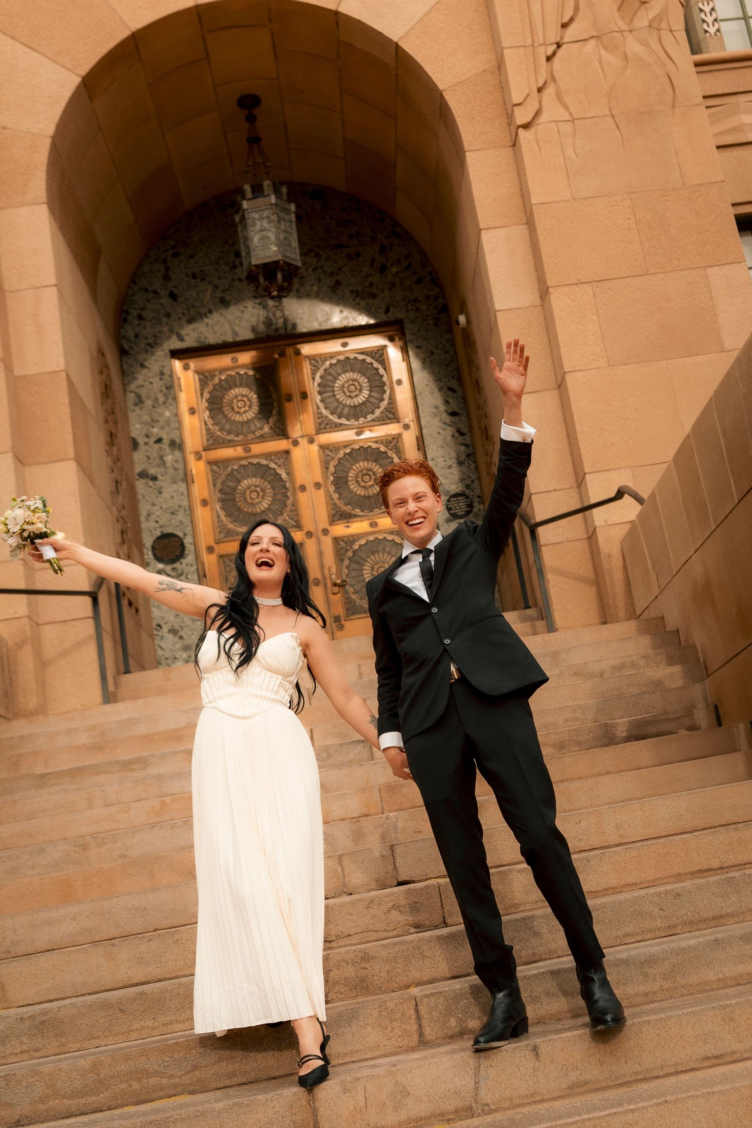 Joyful newlyweds celebrate on church steps, bride in white gown holding bouquet, groom in black suit, both raising arms.