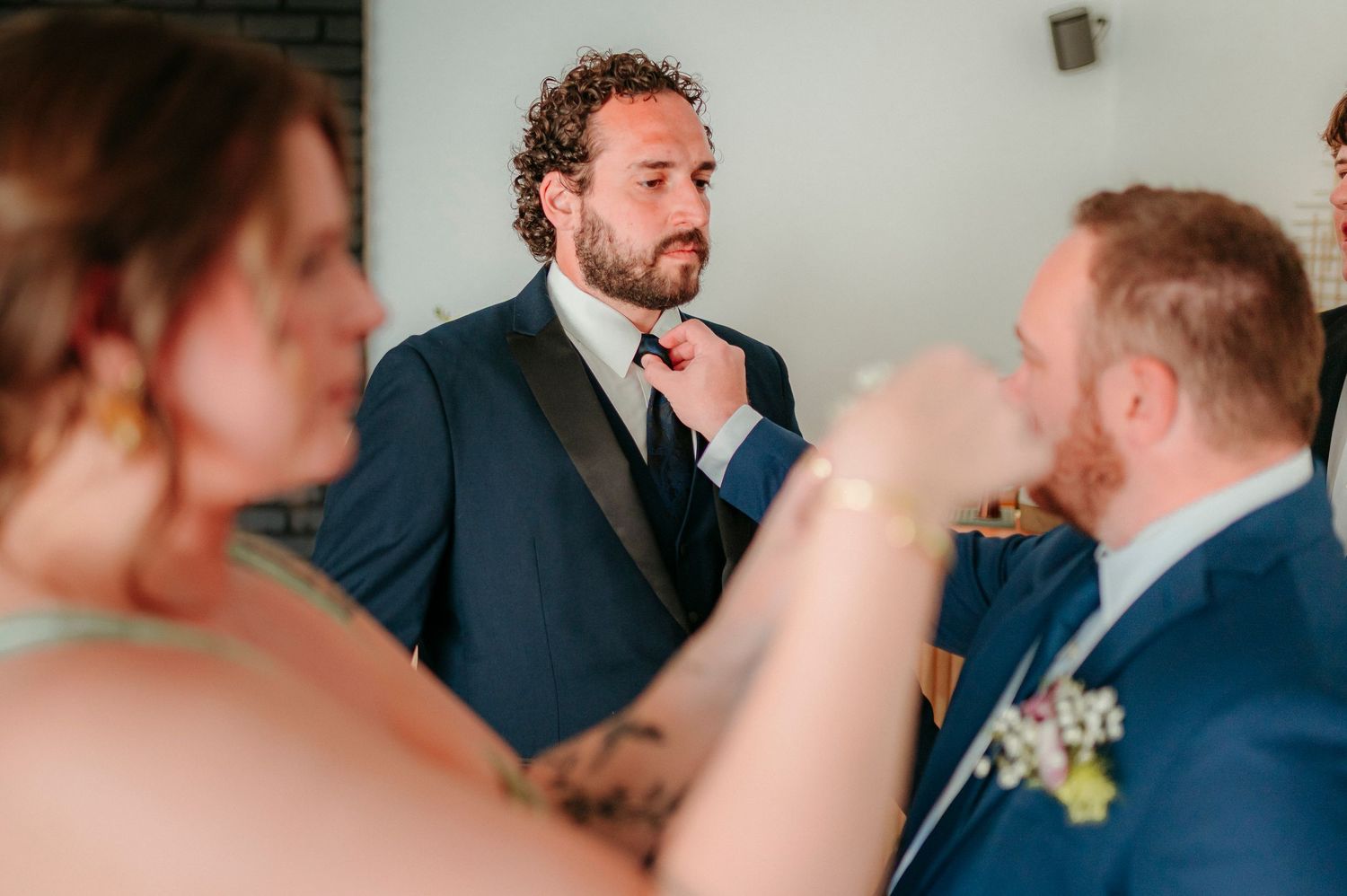 Groom in navy tuxedo adjusting his bow tie with groomsmen nearby during wedding preparations.