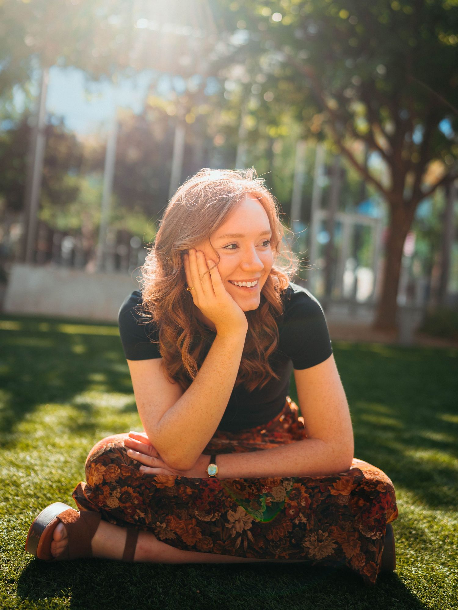 Smiling redhead woman sitting cross-legged on grass in sunny park setting.
