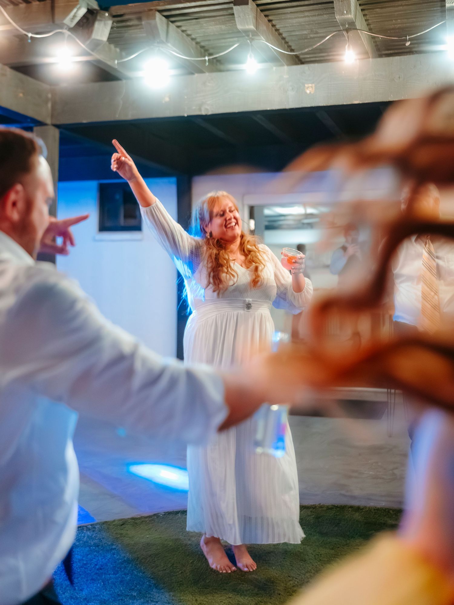 Woman in white dress dancing joyfully with arm raised at an indoor party with string lights.