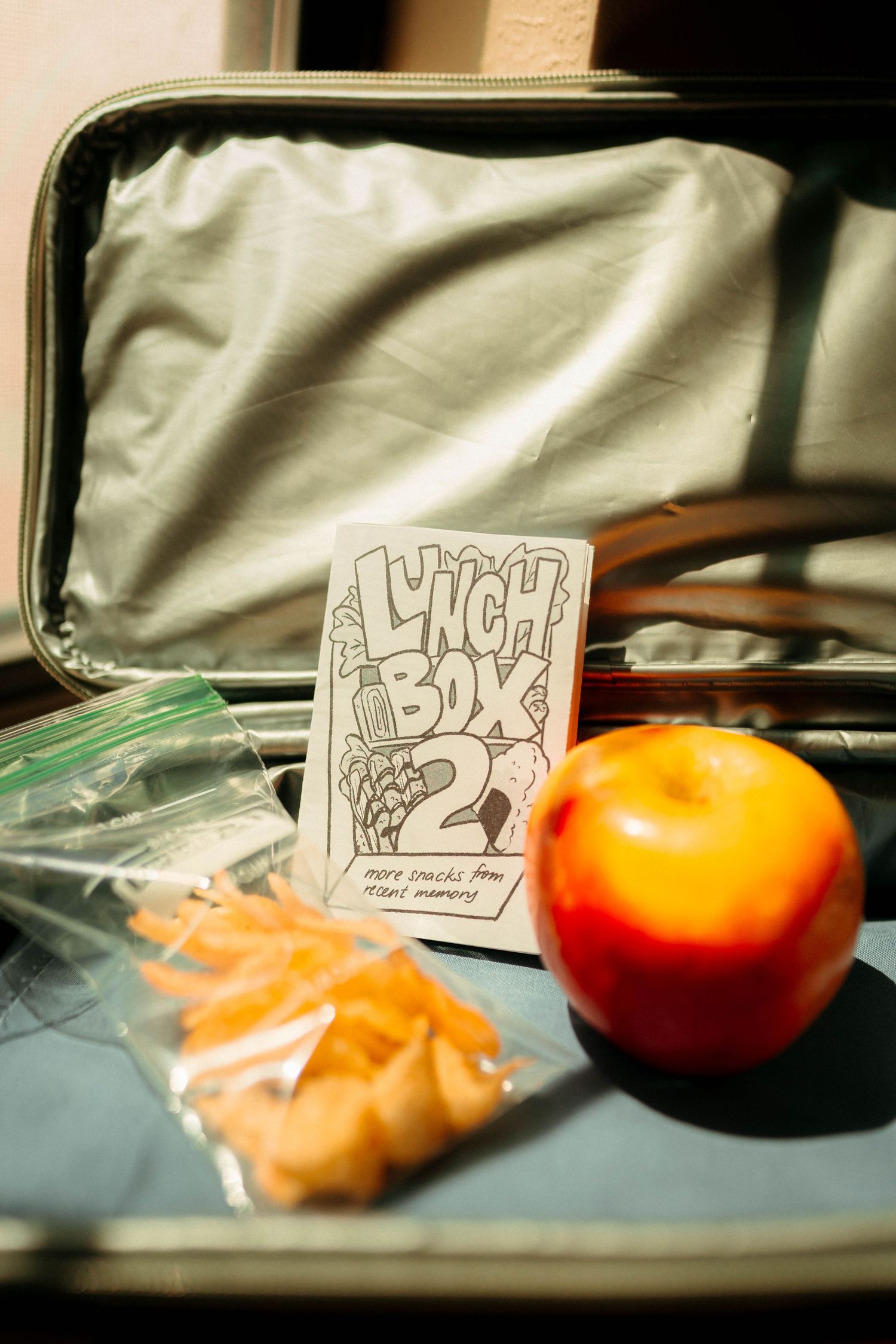 Ash Does Comics' Lunch Box 2 zine inside an open suitcase with an apple and bag of snacks.