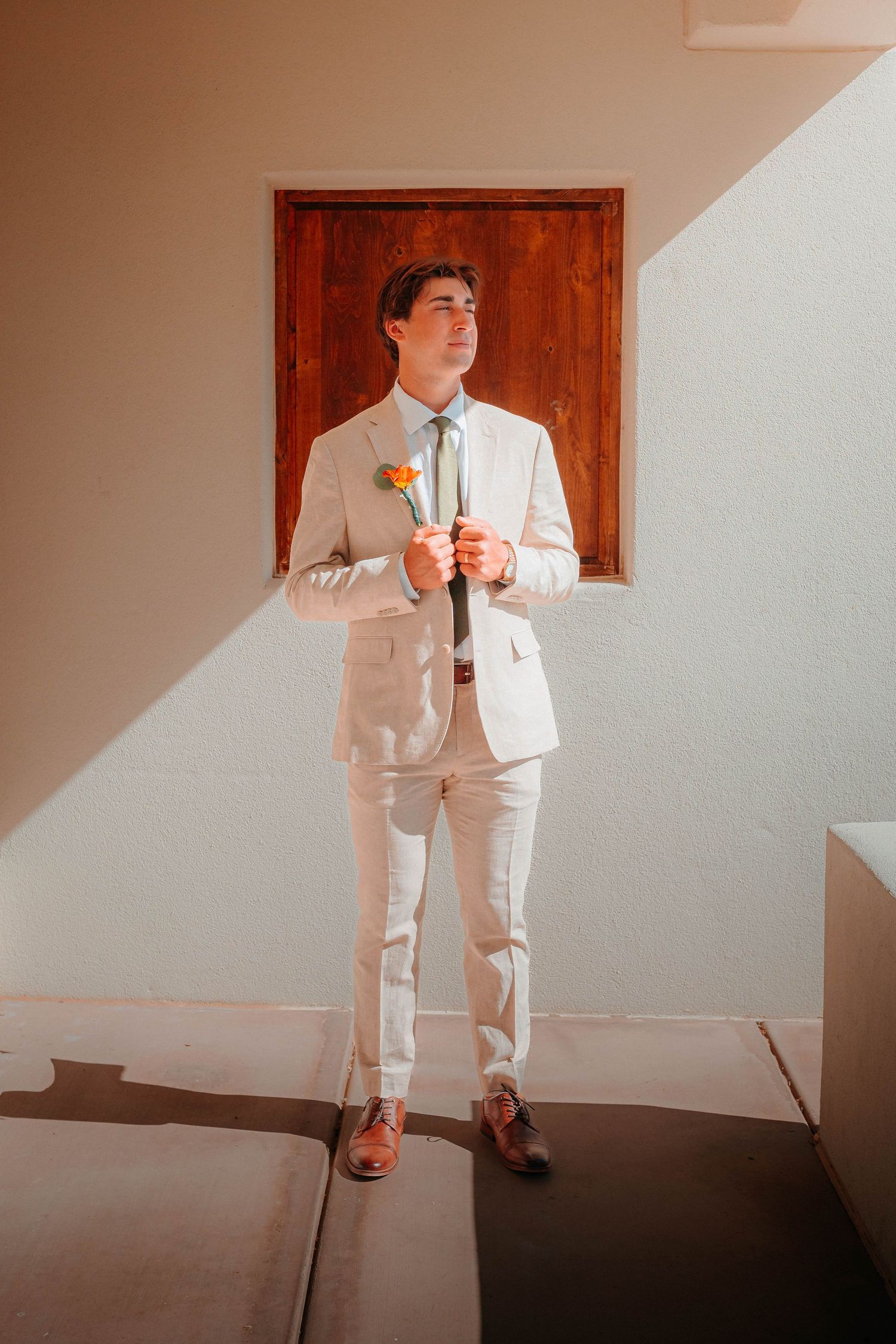 Dapper groom in beige suit holds orange boutonniere while standing in dramatic natural light against white wall.