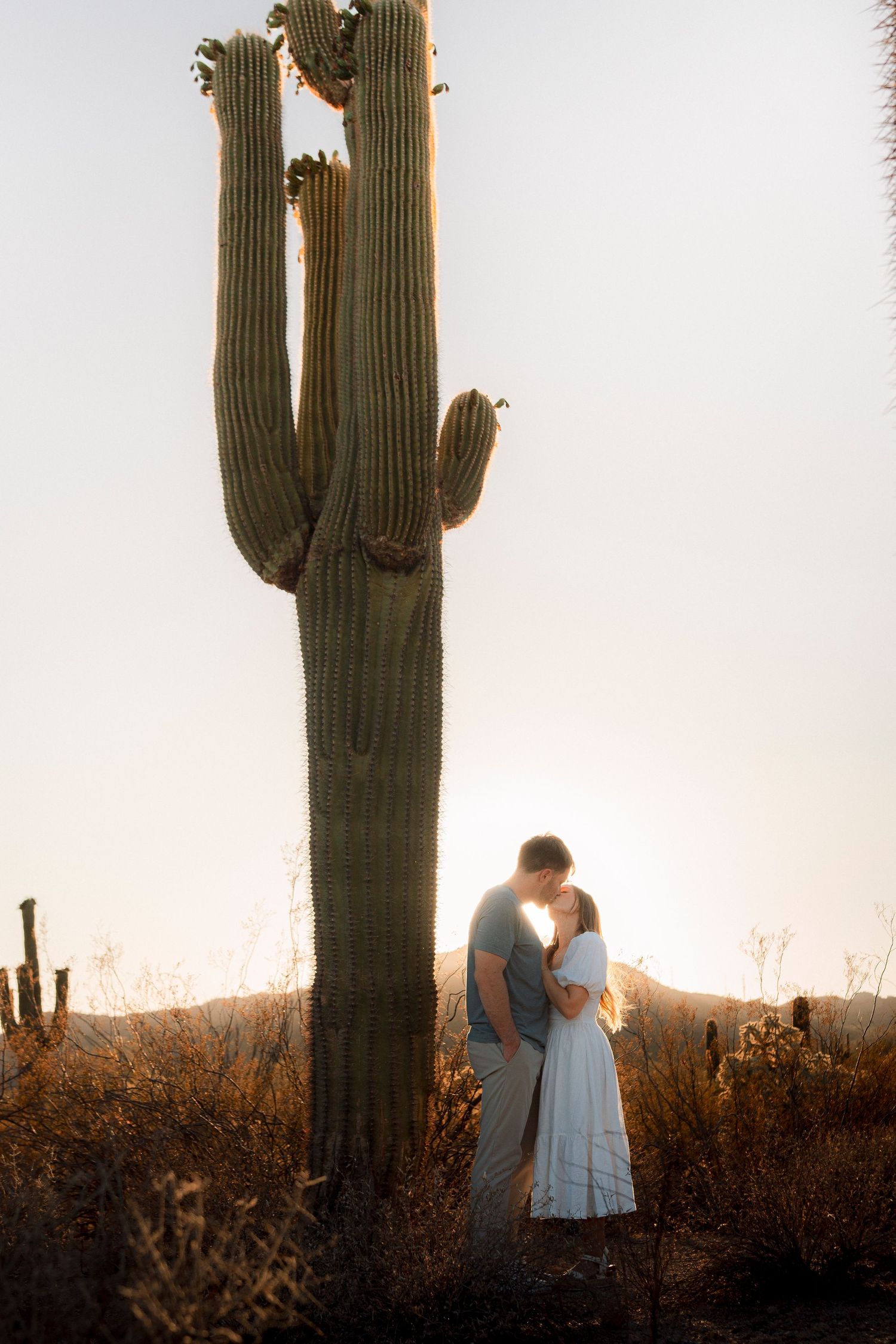 Couple kissing at sunset beside a towering saguaro cactus in the Arizona desert.
