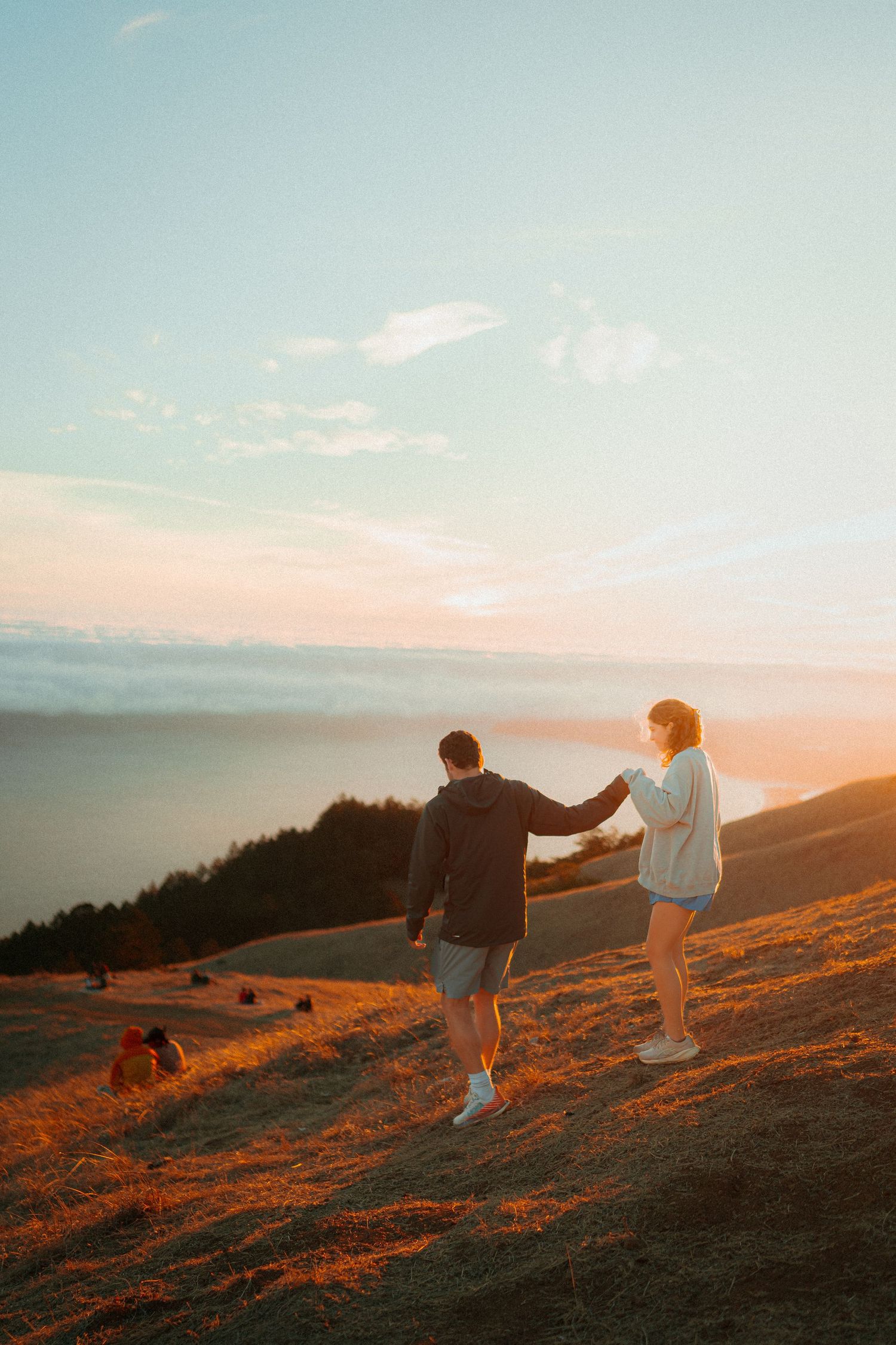 Couple holding hands on a golden hillside at sunset with ocean views in the background.