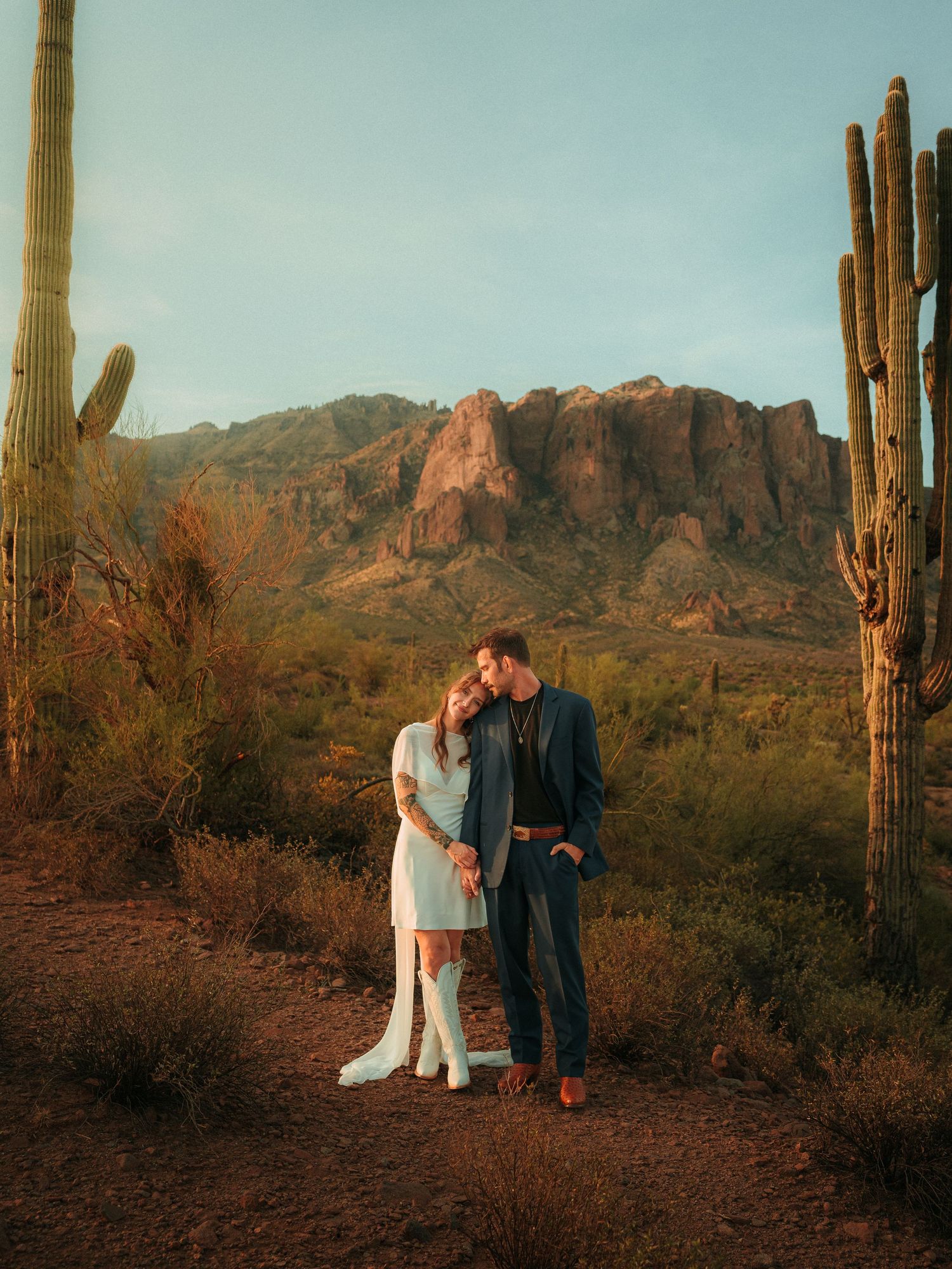 Couple shares a romantic kiss surrounded by saguaro cacti with Superstition Mountains at golden hour in Arizona desert.
