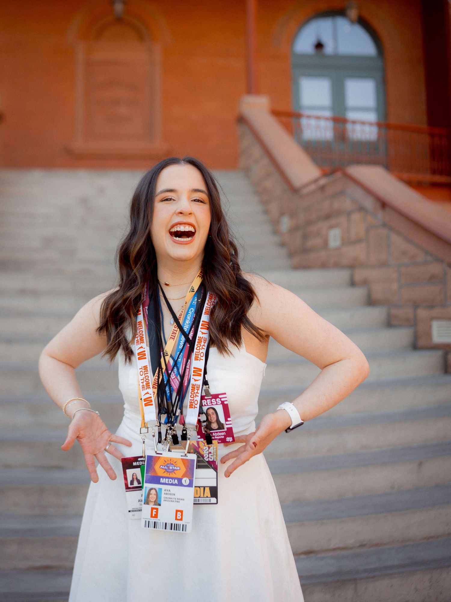 Laughing young woman in white dress poses with hands on hips wearing lanyards on steps.