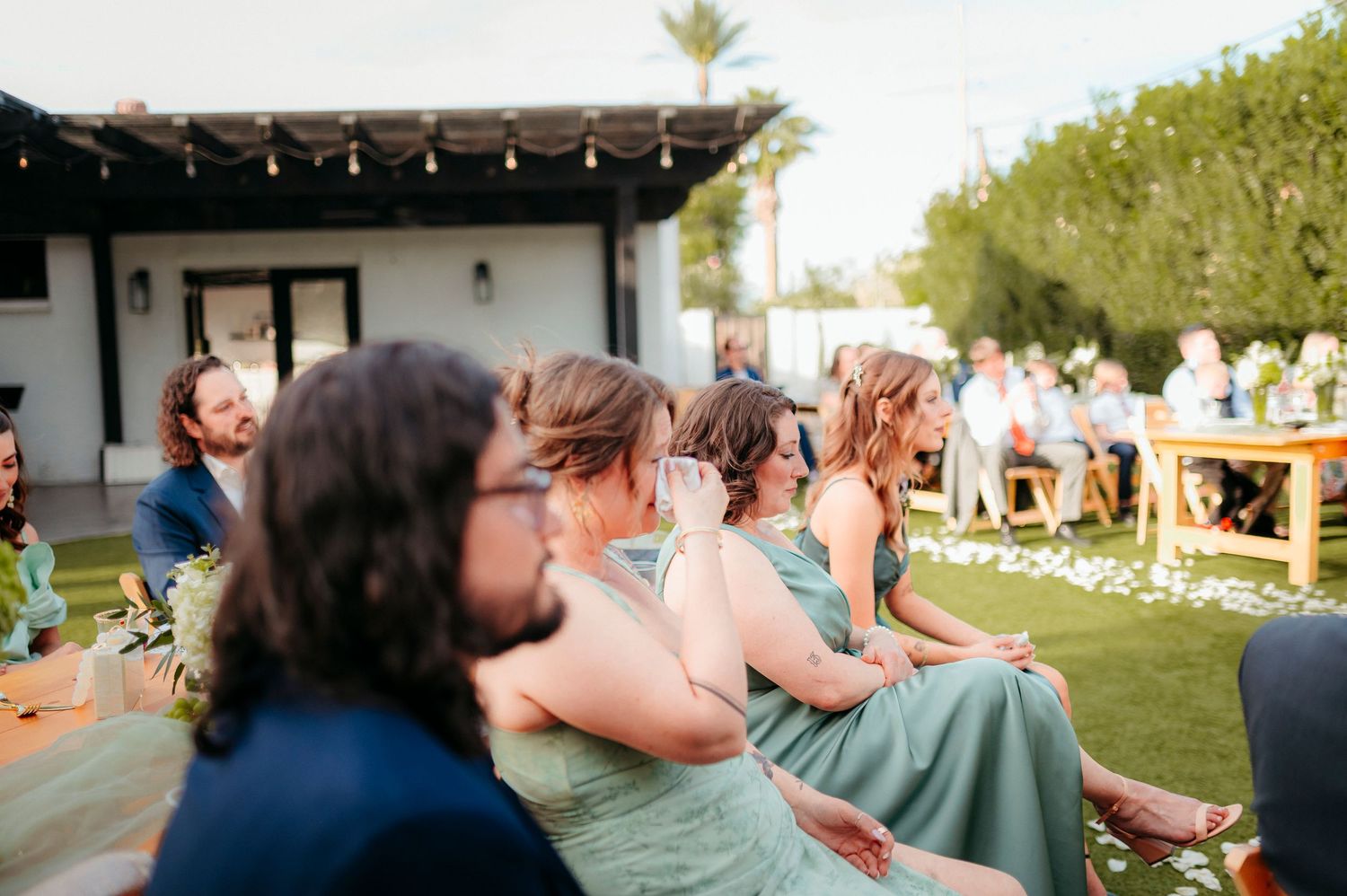 Guests in sage green dresses sit in rows during an outdoor wedding ceremony on a sunny day.