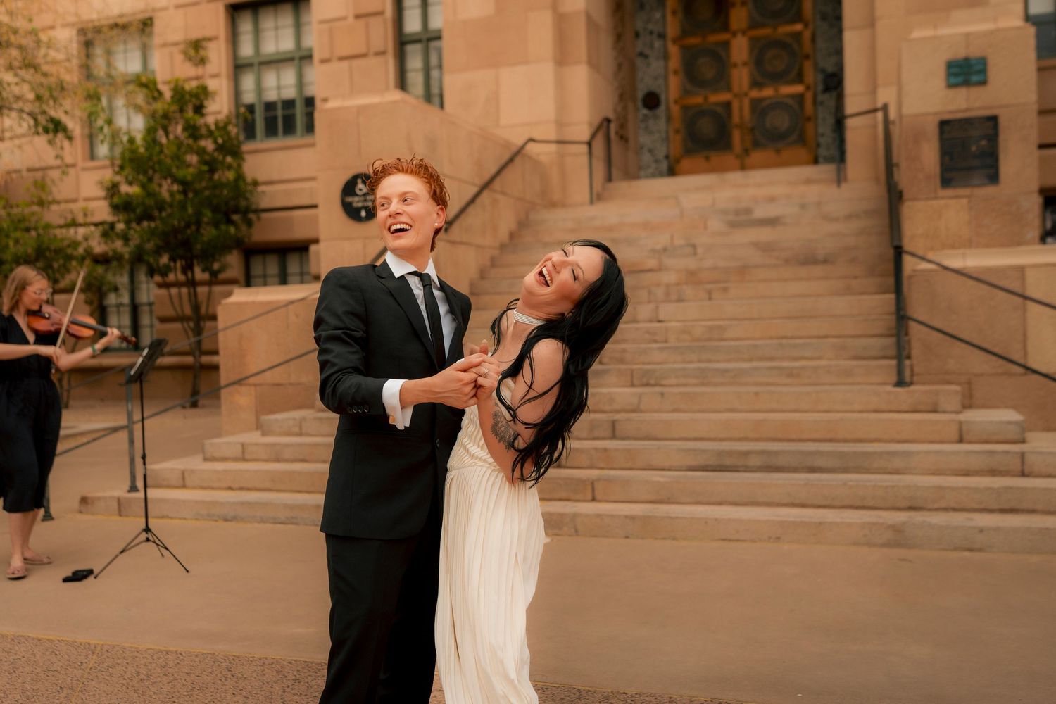 Couple dancing joyfully outside a stone building, bride in white gown, groom in black suit, violinist performing in background.