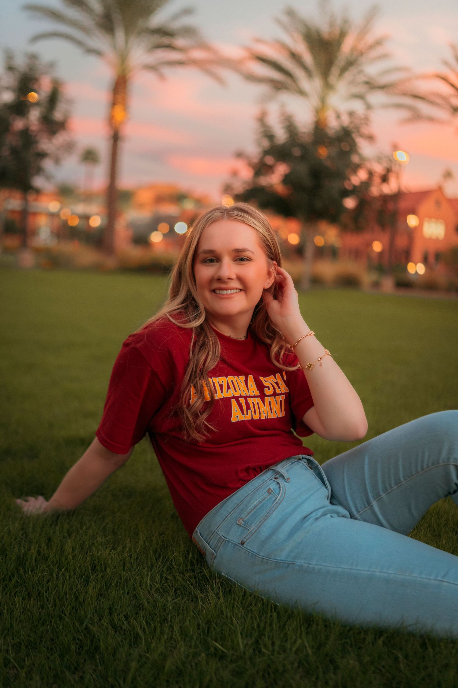 Smiling young woman in Arizona State Alumni shirt relaxing on grass at colorful sunset campus.