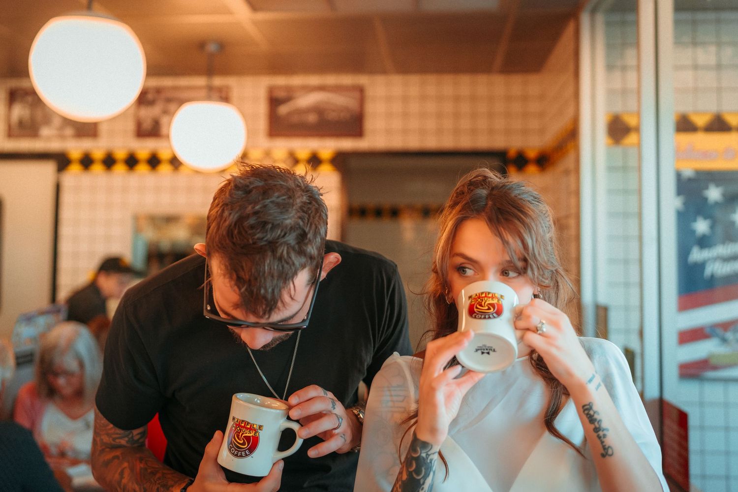A tattooed couple drinking coffee from branded mugs at a retro American diner booth.