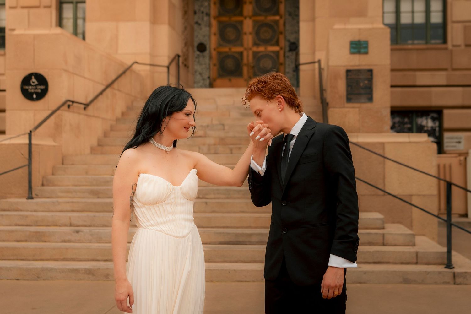 Groom in black suit kisses bride's hand as she smiles, standing before stone steps at an outdoor wedding venue.