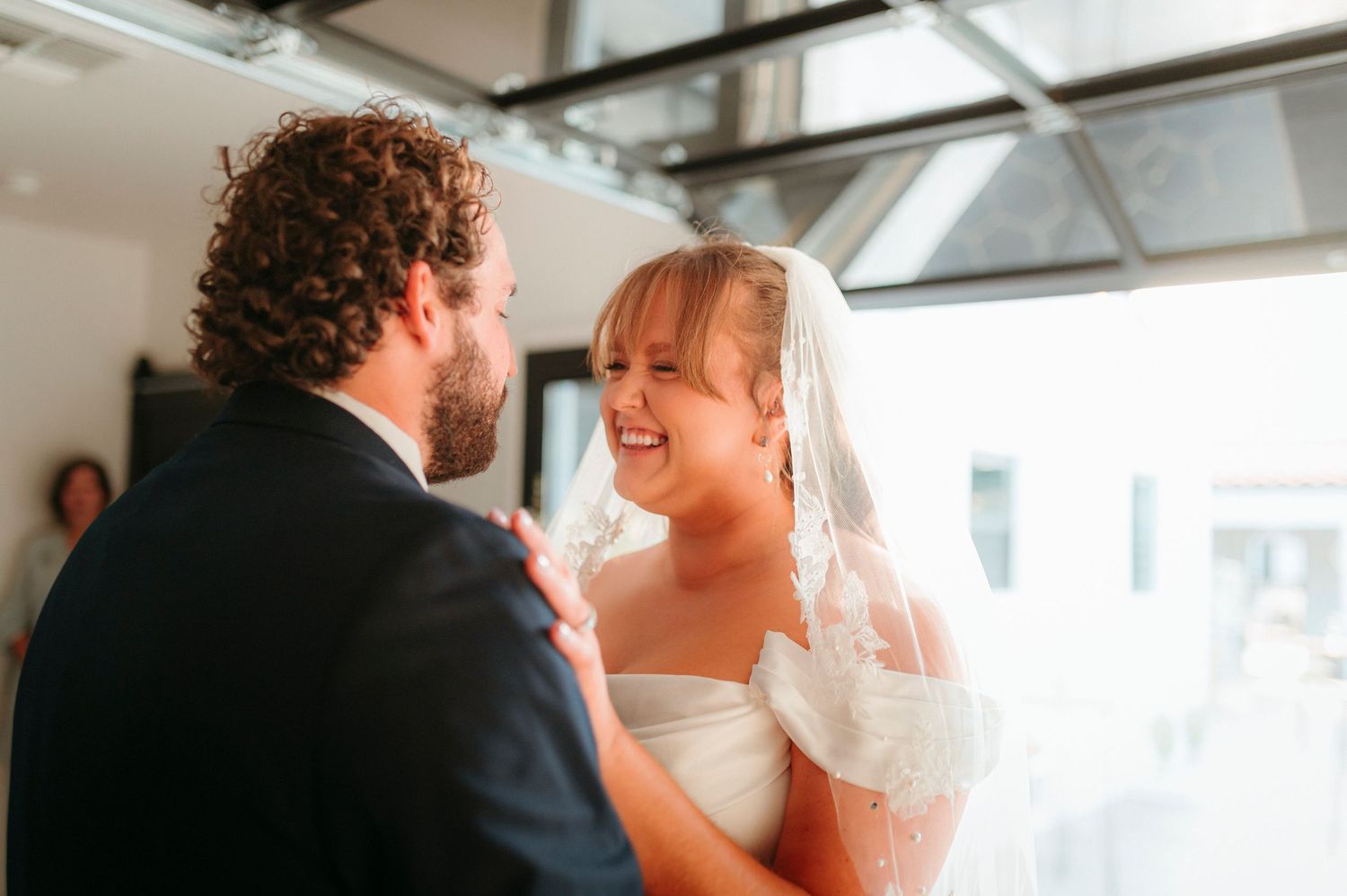 Bride in lace veil laughing joyfully with groom during their wedding day first look moment.