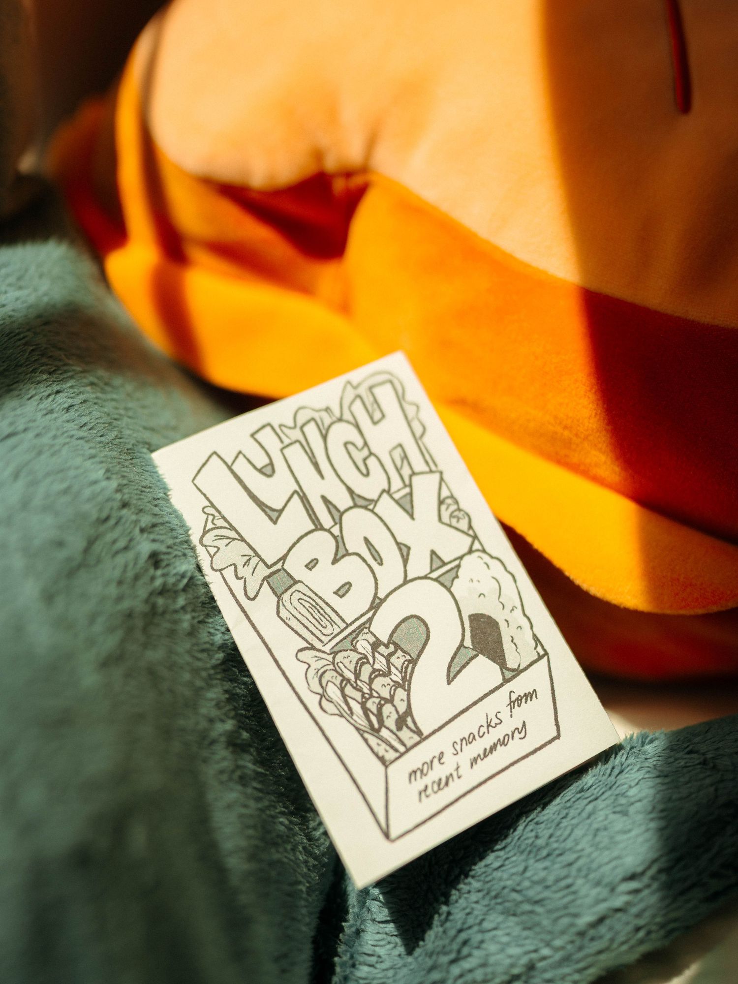 White zine titled Lunch Box 2 resting on orange and teal fabric in soft light.