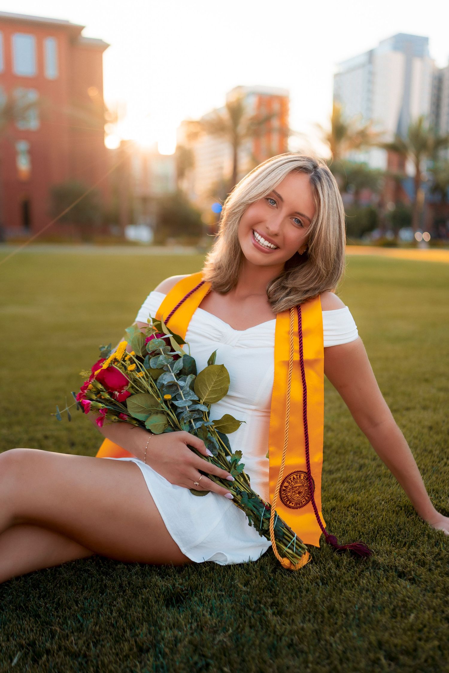 Smiling blonde woman in white dress holds roses wearing gold graduation stole on green lawn.