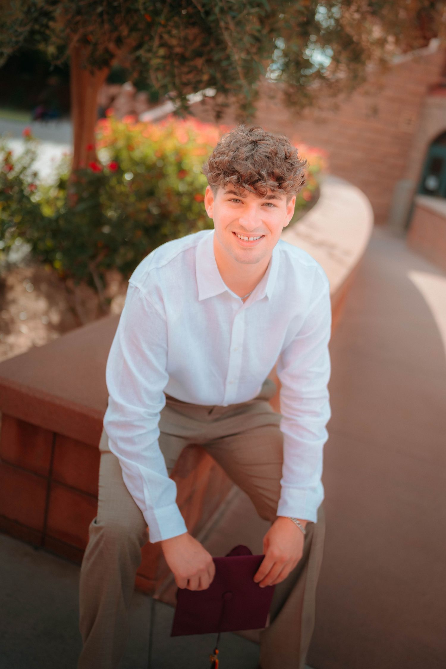Smiling young man in white shirt leans forward holding diploma outdoors near brick wall and greenery.