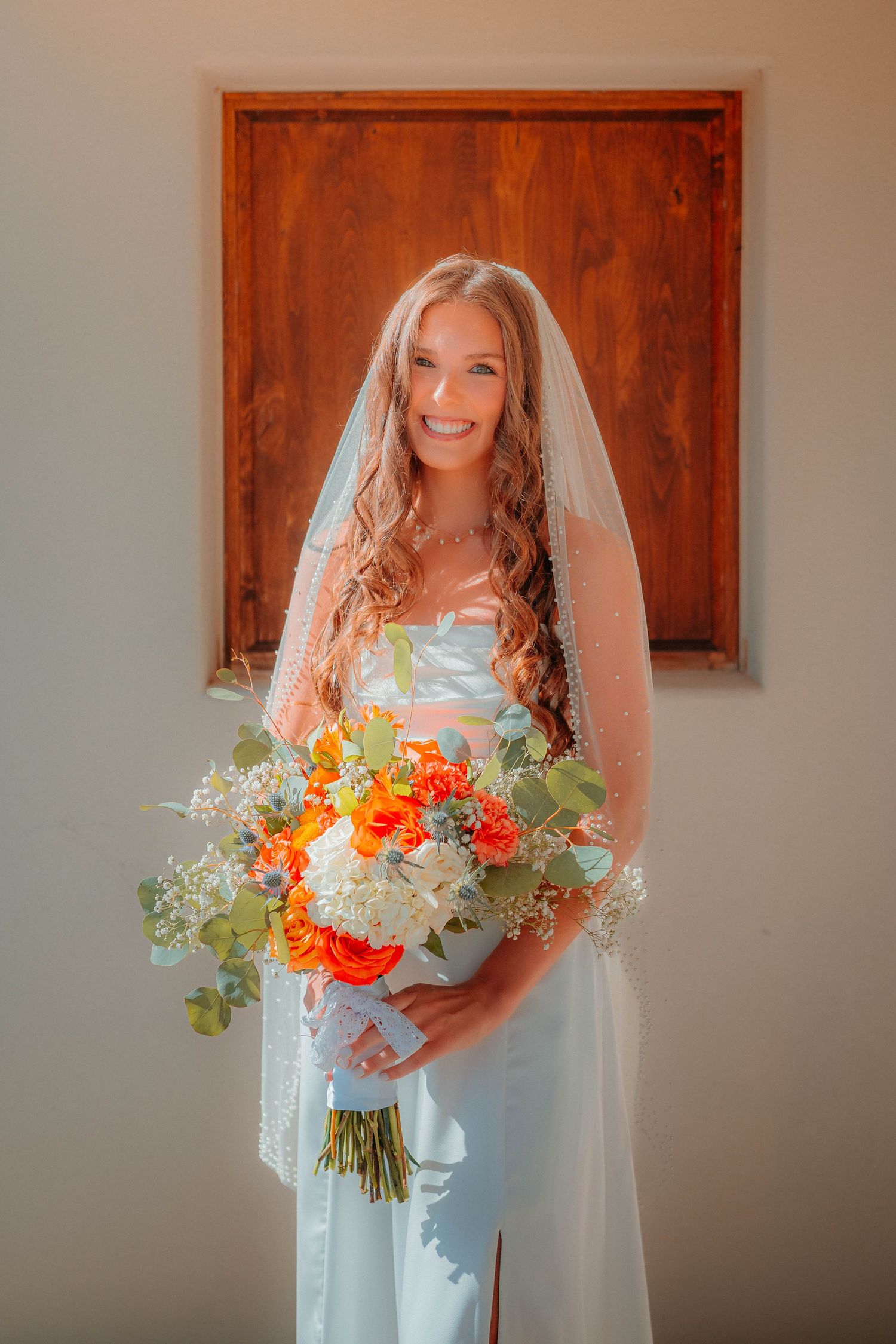 Radiant bride smiles wearing a white gown and veil while holding a lush orange and white floral bouquet.