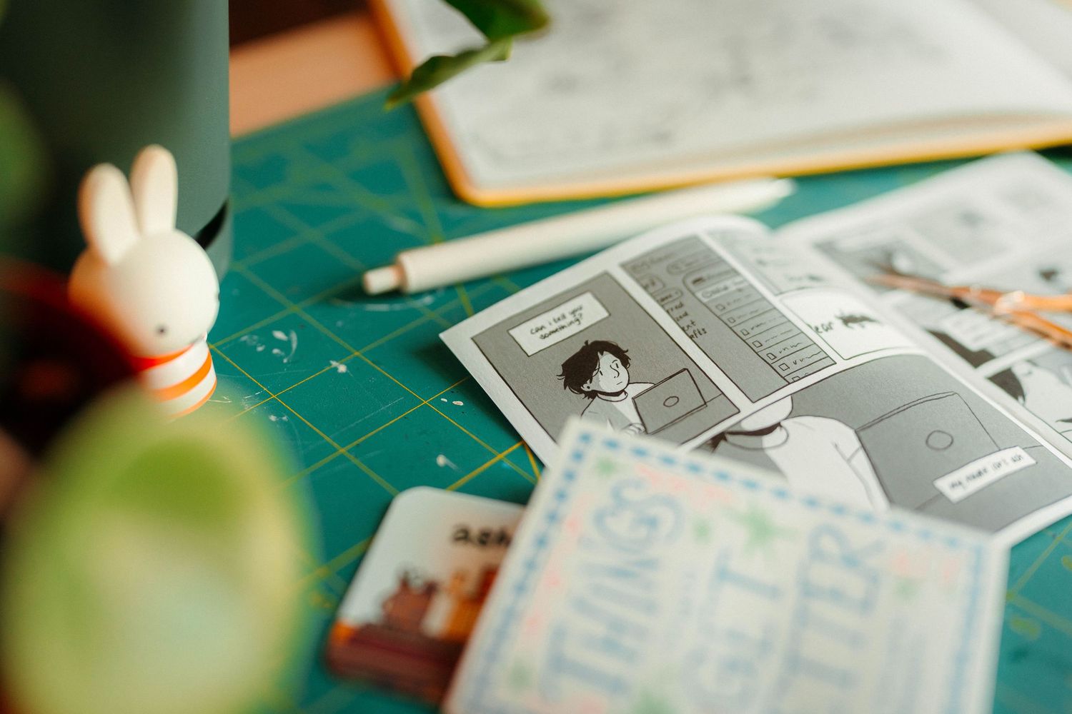 Small comic zine pages spread on green cutting mat with Miffy figurine and plant.