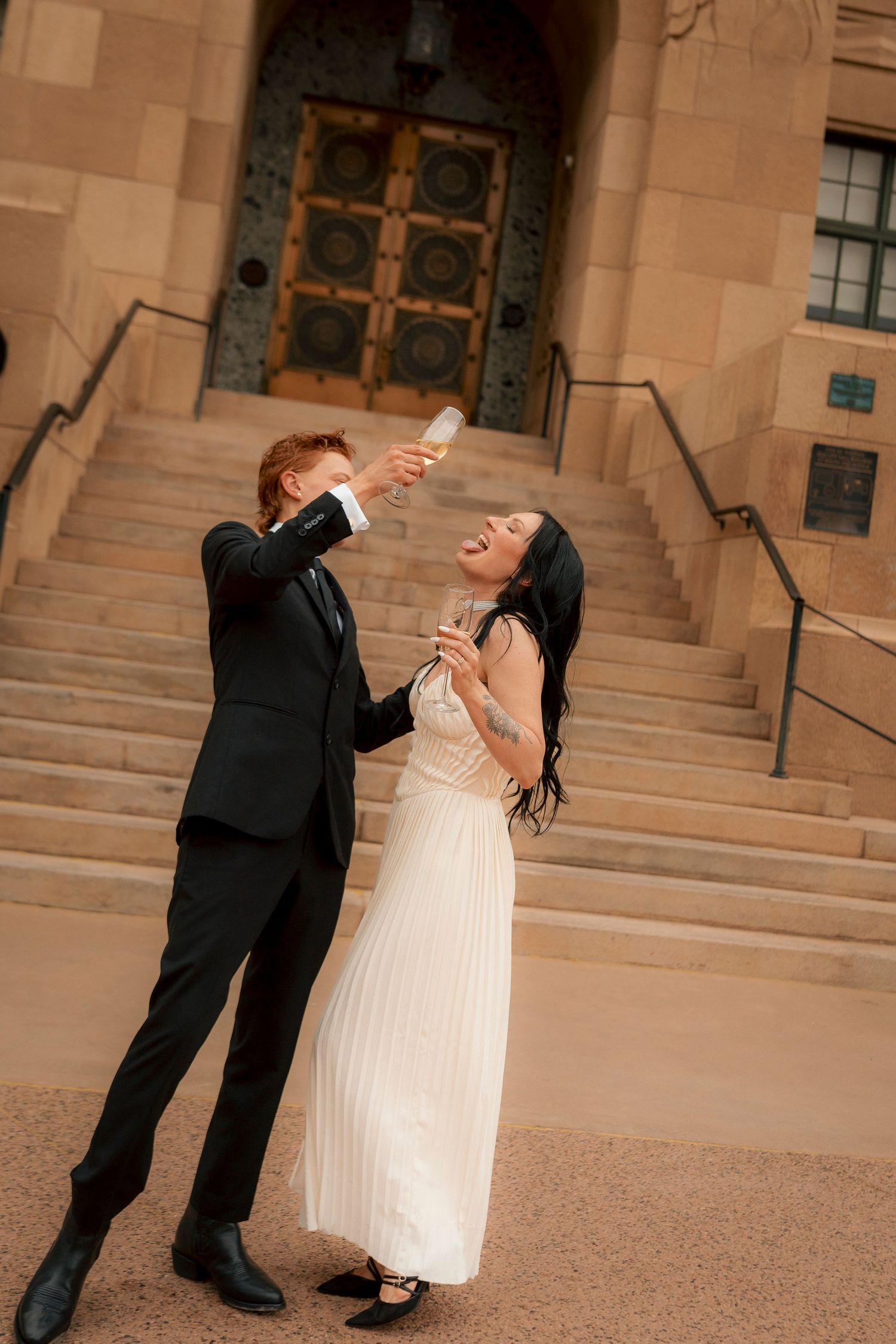Joyful newlyweds toasting with champagne outside a grand stone building, bride in white gown, groom in black suit.