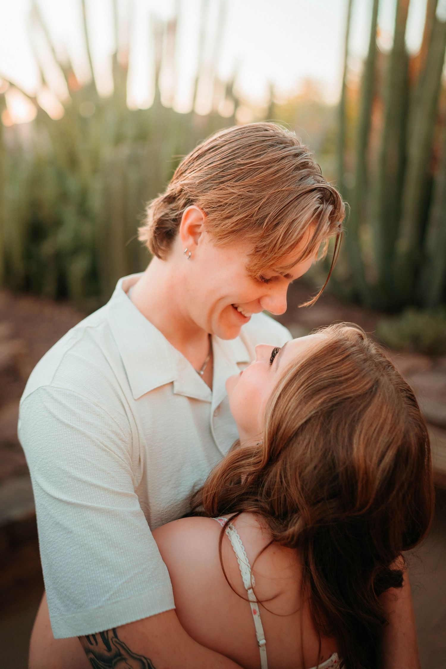 A couple shares a romantic moment, nearly kissing and smiling, surrounded by tall cacti in warm golden light.