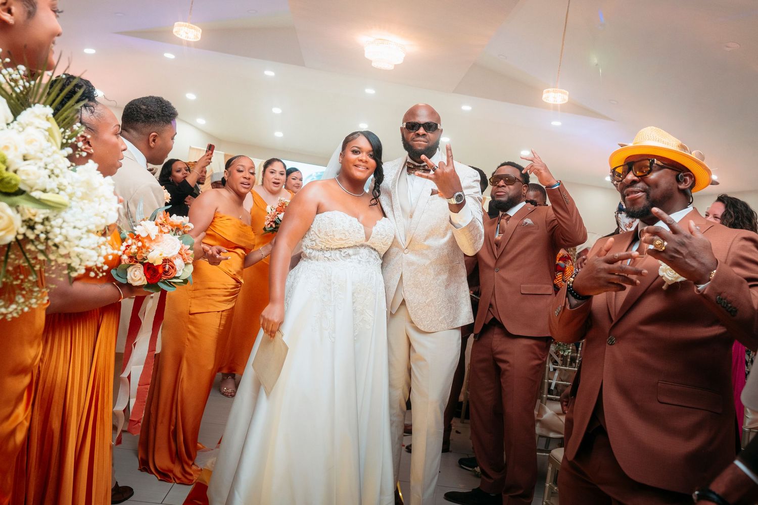 Bride and groom joyfully enter wedding reception surrounded by orange bridesmaids and brown groomsmen.