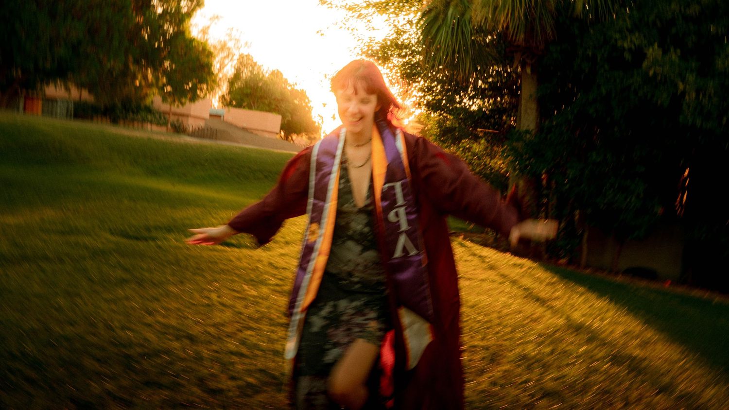 Graduate in maroon cap and gown with sash running on green grass during golden sunset.