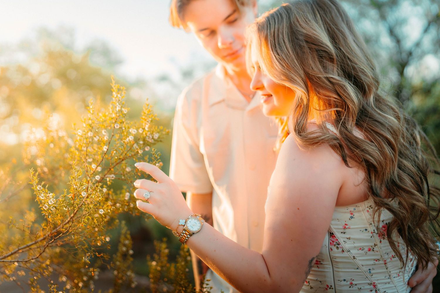 A smiling couple examines a yellow flowering bush together in warm golden sunlight outdoors.