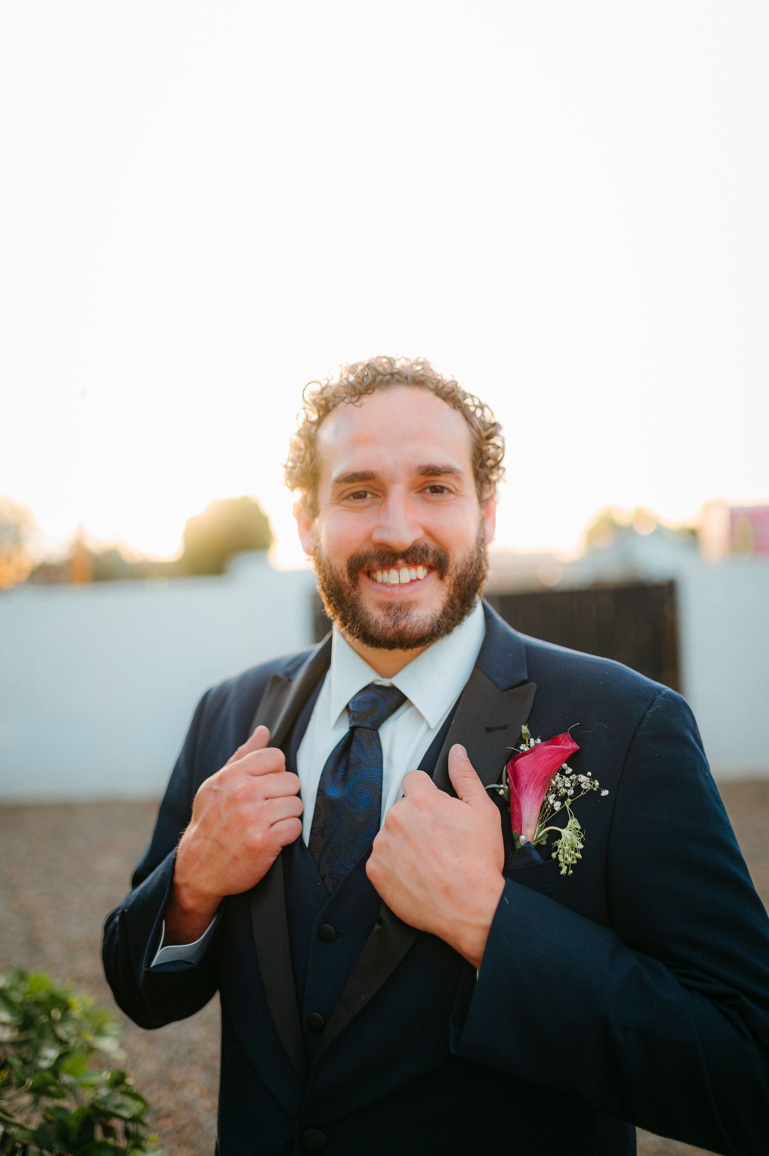 Smiling groom in navy tuxedo with pink boutonniere adjusting lapels outdoors at golden hour.