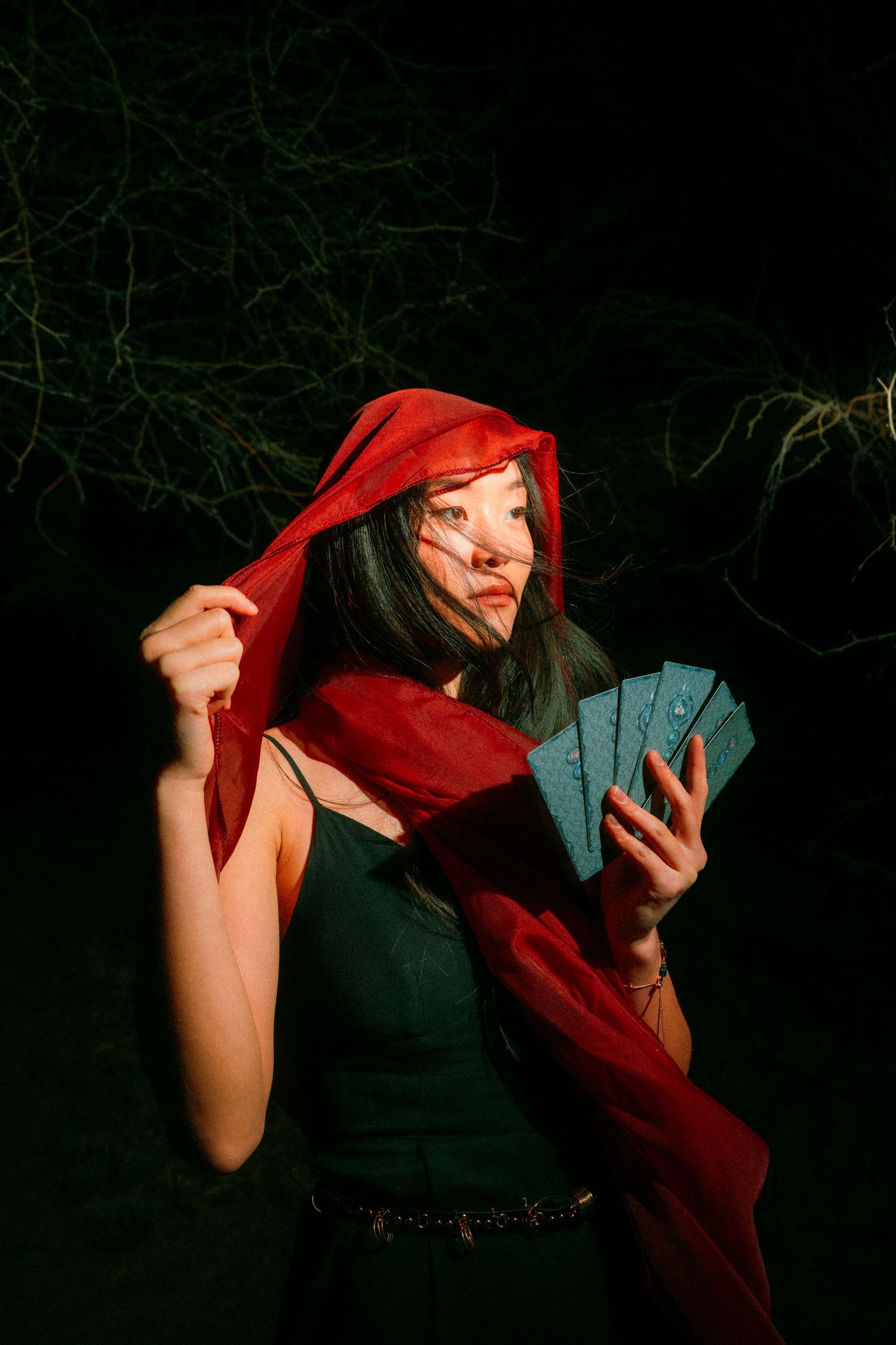 Mysterious woman in red hood holding tarot cards in a dark forest night setting.