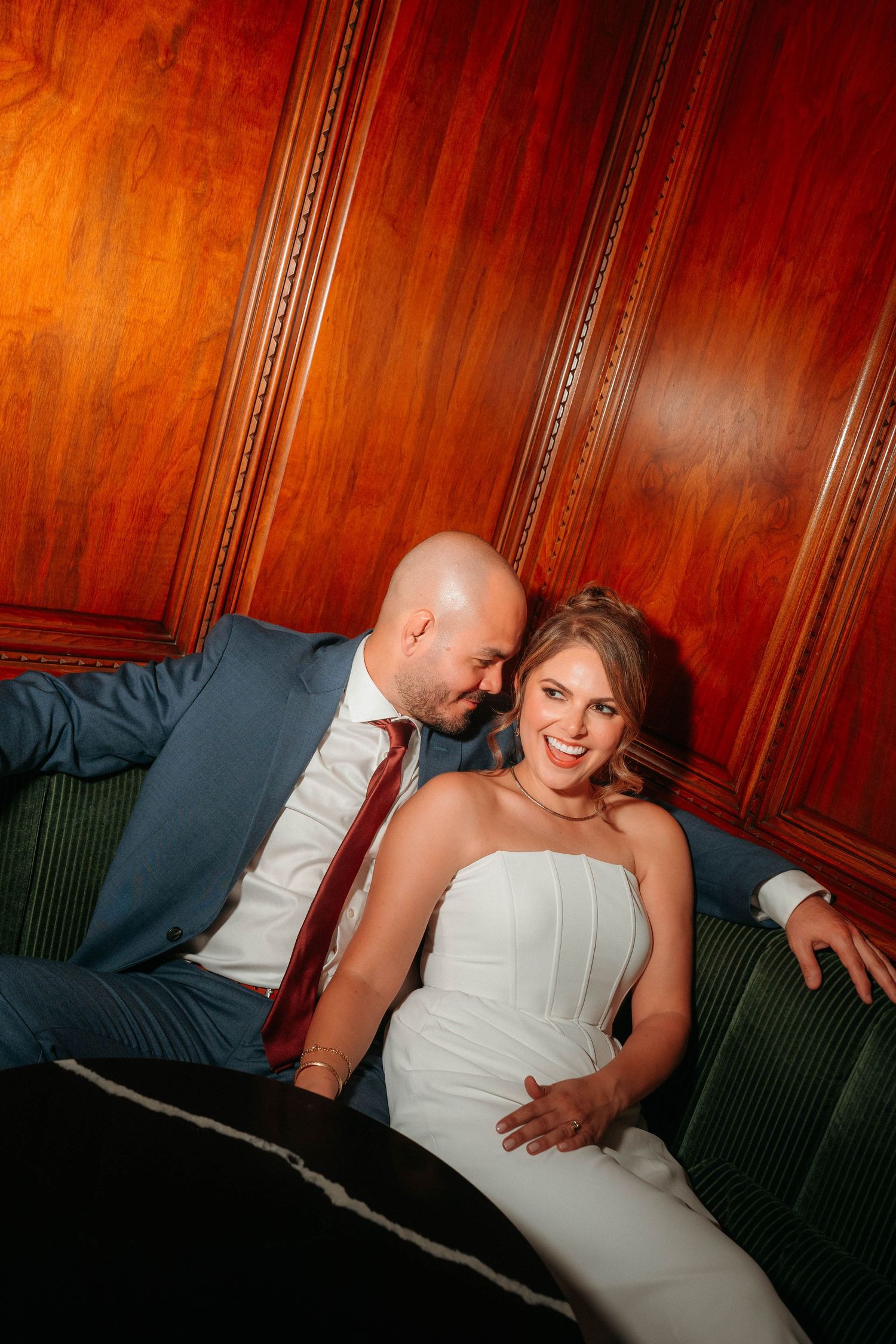 Smiling bride in white gown and groom in blue suit relax on a green velvet sofa.