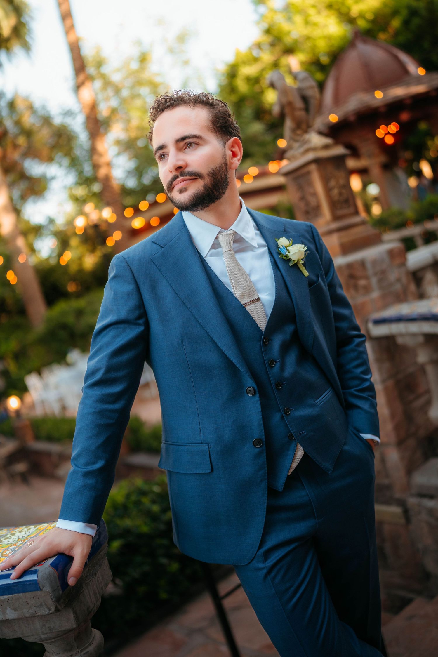 Handsome groom in a navy blue three-piece suit with boutonniere posing outdoors at a garden wedding venue.