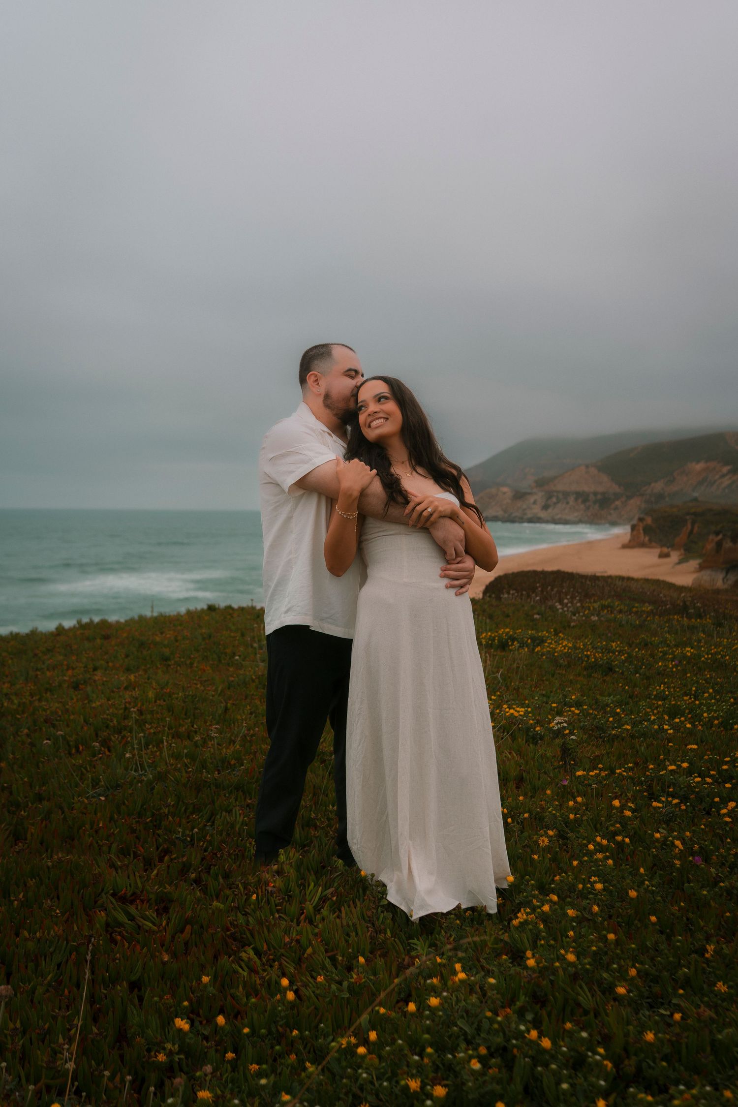 Couple embracing on a coastal cliff with ocean views and wildflowers during an engagement photo session.