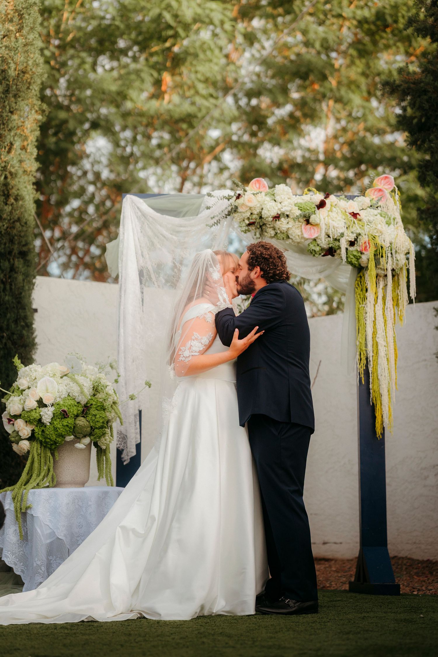 Bride and groom share their first kiss at an outdoor wedding ceremony beneath a floral arch with white and pink blooms.
