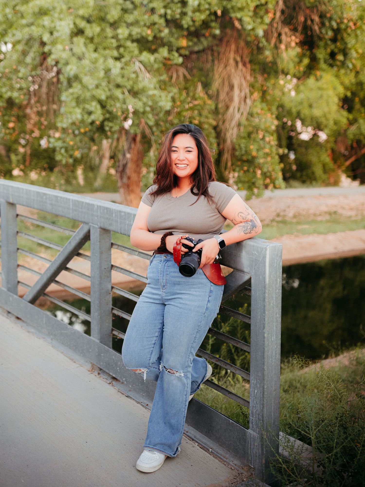 Smiling female photographer leaning on bridge railing holding camera in lush green park setting.