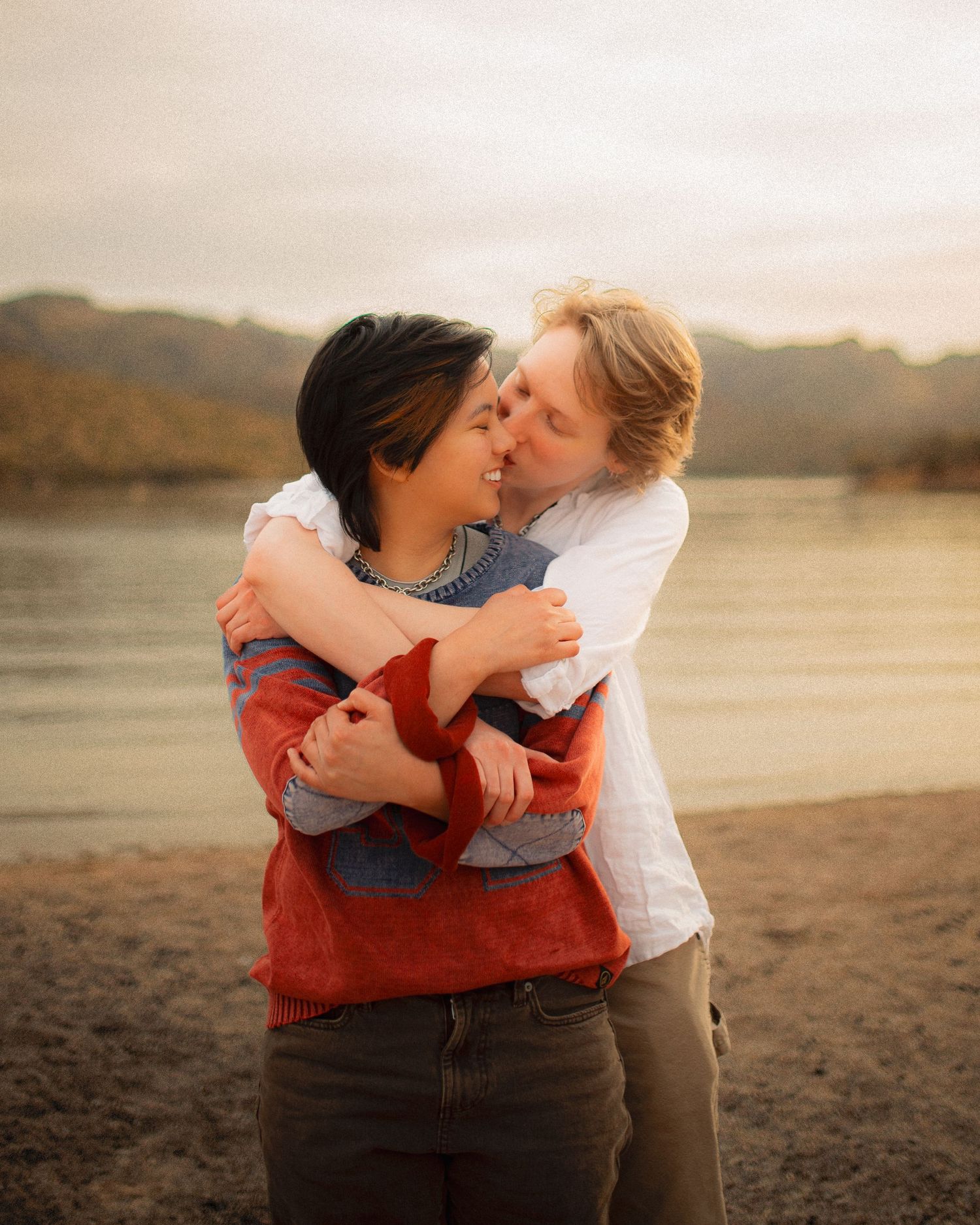 Two people sharing a tender embrace and kiss by a lakeside with mountain backdrop at dusk.