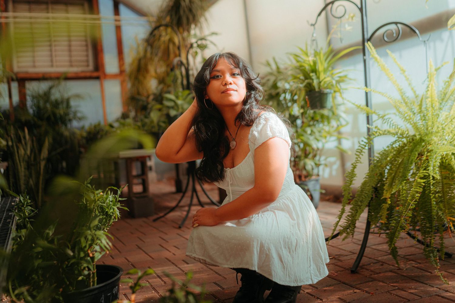 Young woman in light blue dress sits among lush green plants in sunlit greenhouse setting.