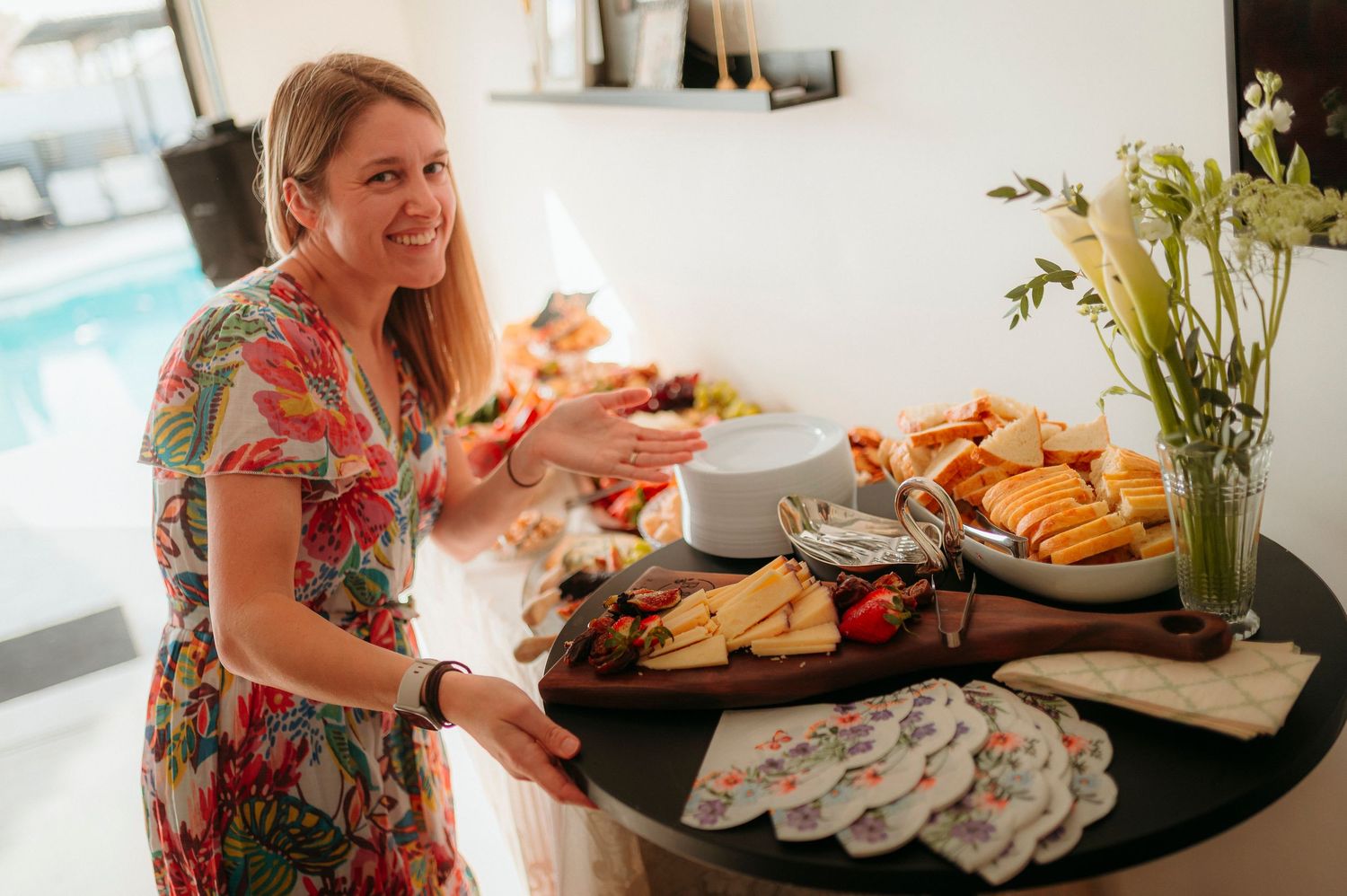 Woman in floral dress smiling while arranging a charcuterie board with cheese, fruits, and snacks near a pool.