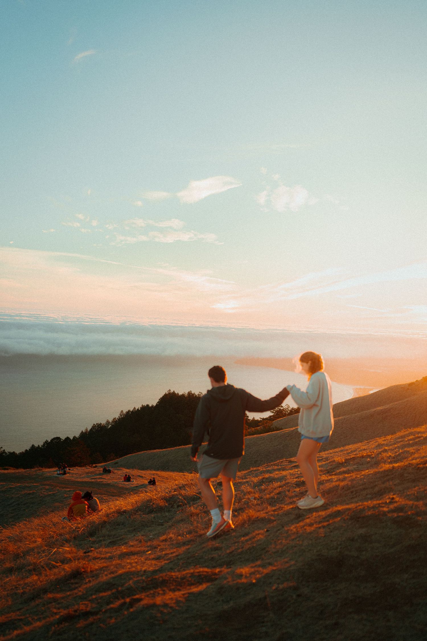Couple holding hands on a golden hillside at sunset with ocean views and glowing sky in the background.
