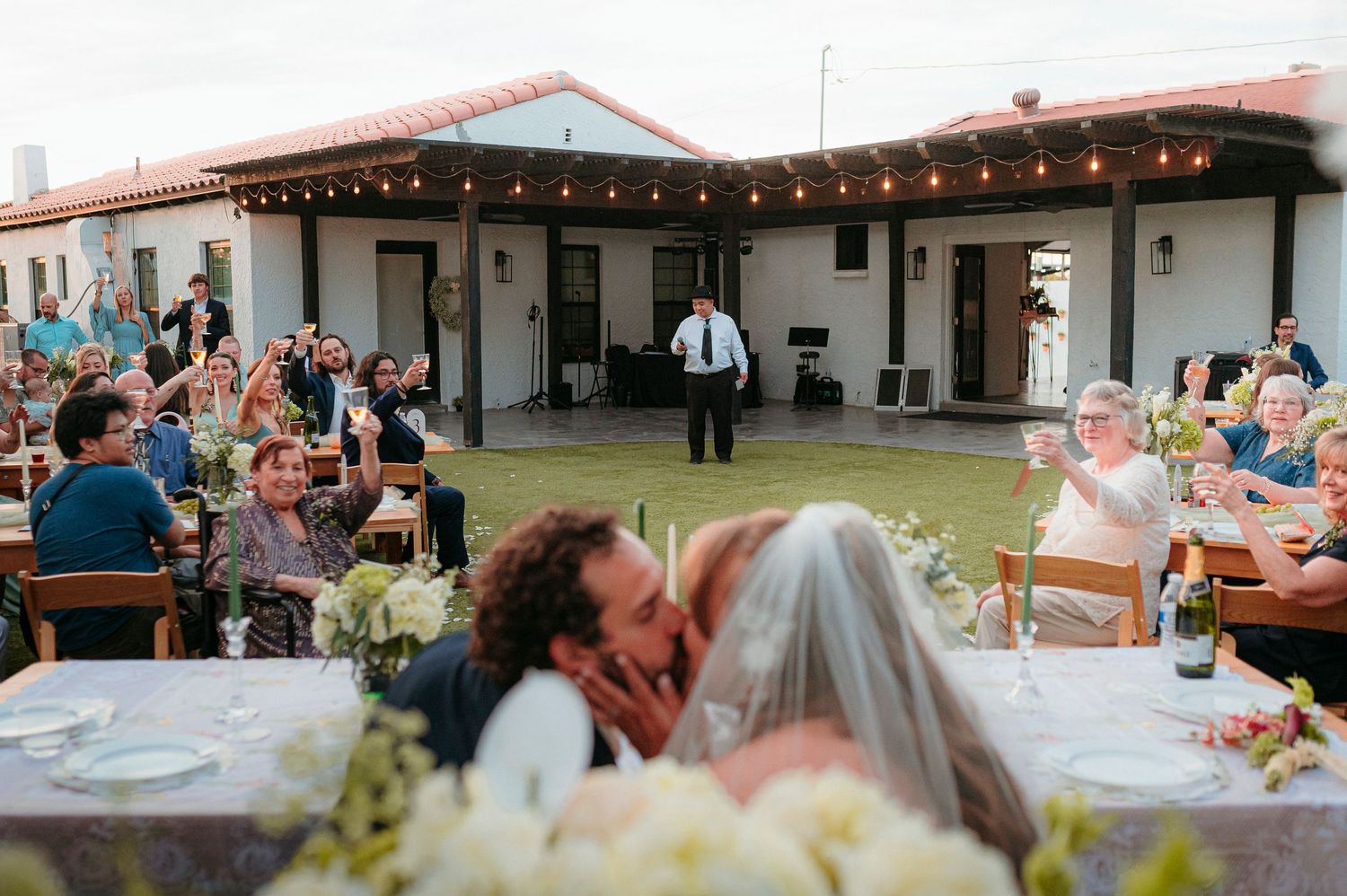 Wedding couple sharing kiss at outdoor reception while guests celebrate under string lights at sunset.