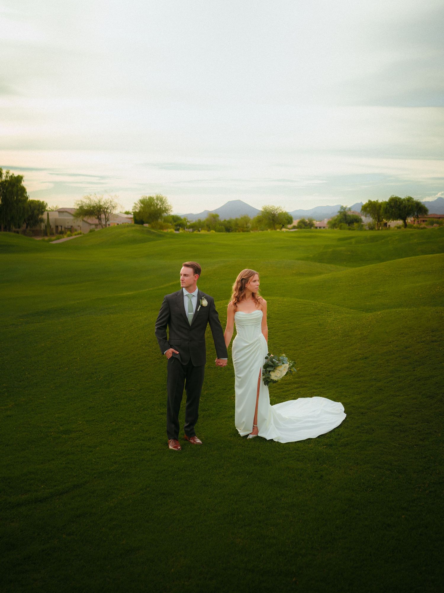 Bride and groom stand on lush green golf course with mountain backdrop at sunset wedding in Arizona.