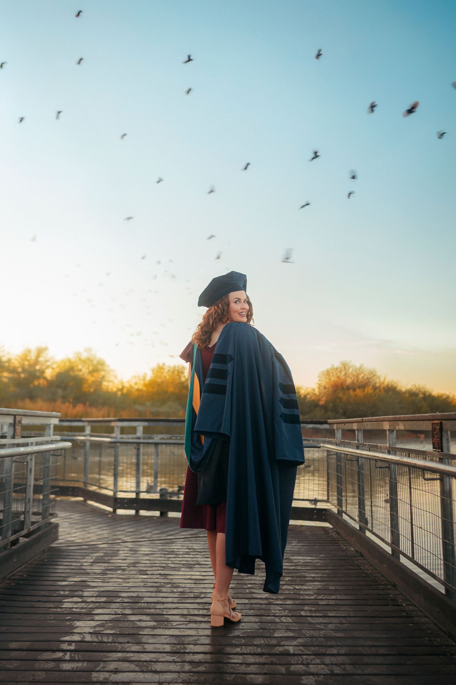 A doctoral graduate in navy regalia stands on a bridge with birds flying overhead.