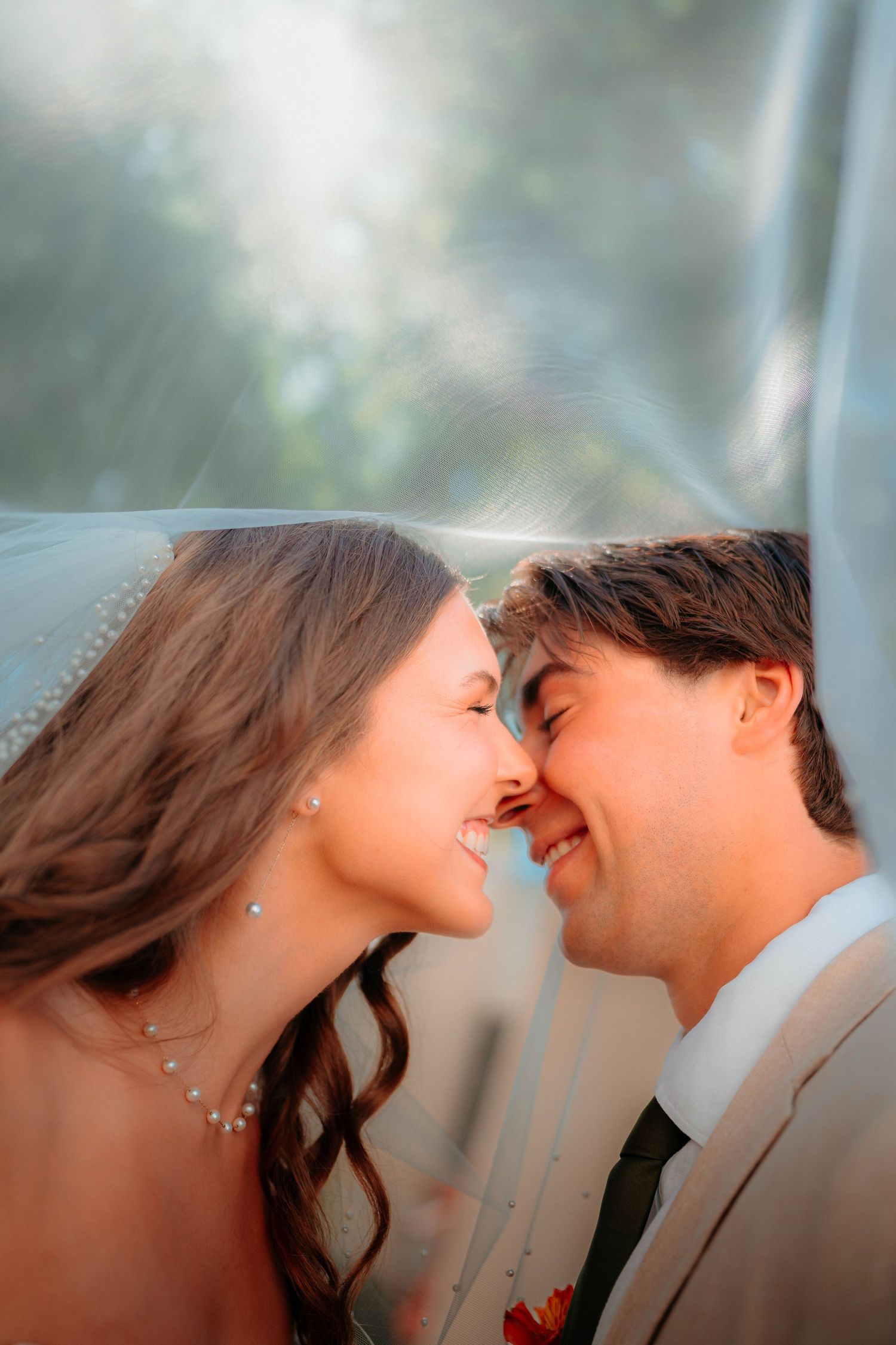 Bride and groom share a romantic nose-to-nose moment under a flowing white veil in soft natural sunlight.