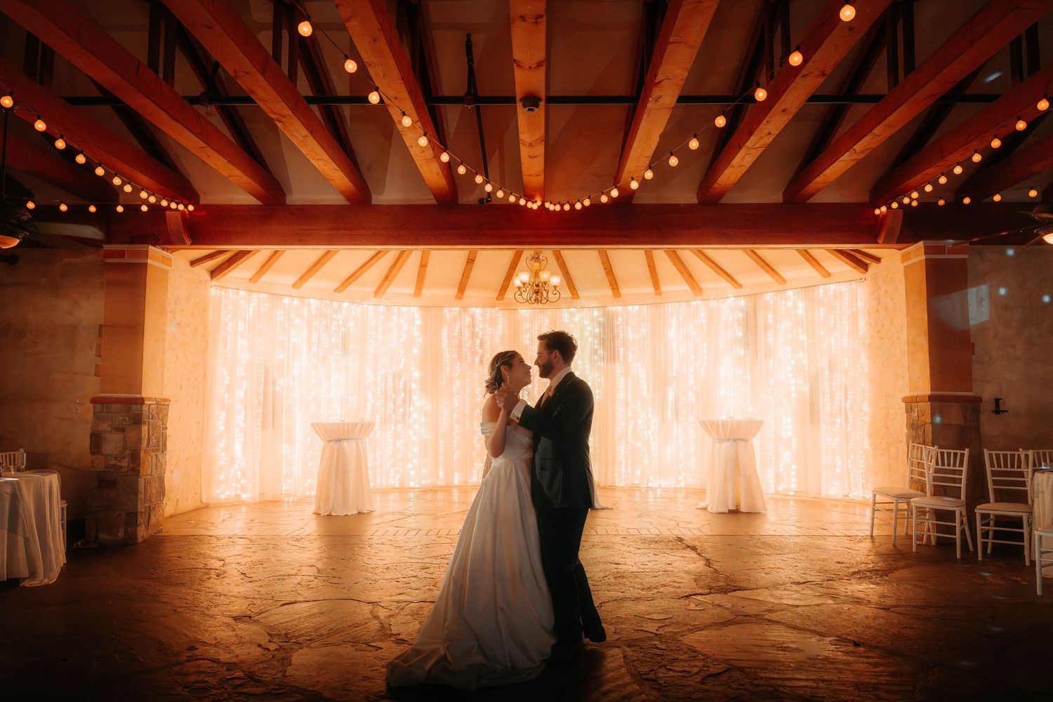 Romantic first dance of bride and groom in a warmly lit rustic venue with fairy lights and string lights backdrop.
