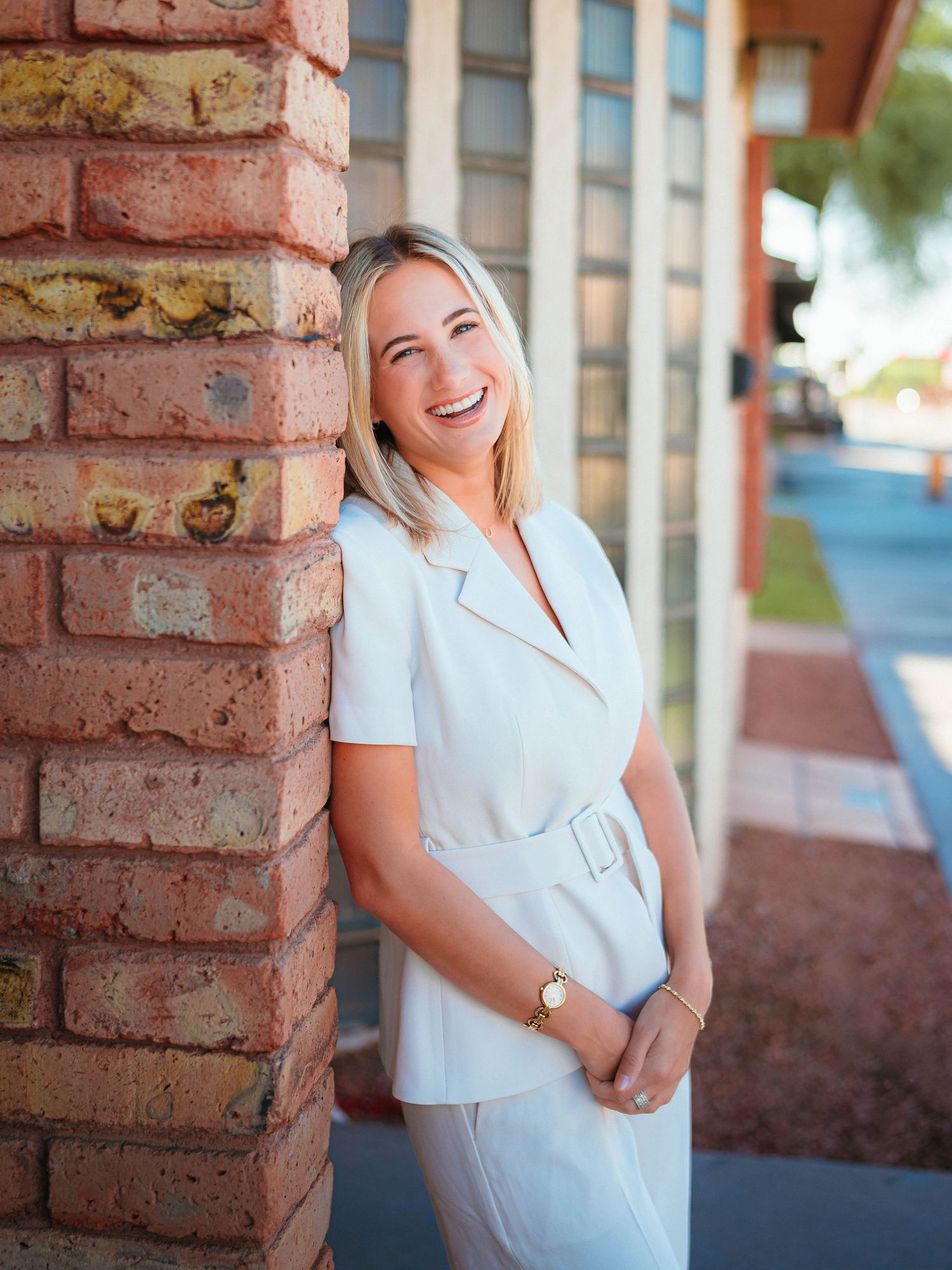 Smiling blonde woman in white outfit leaning against brick pillar in warm light.