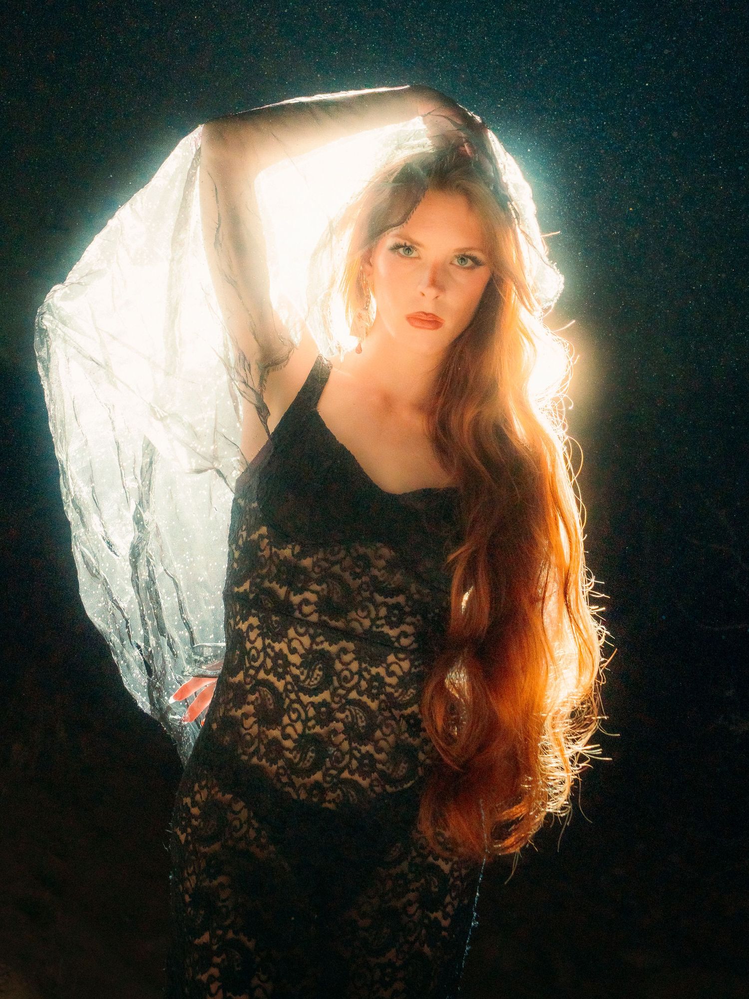Redhead woman in black lace dress holding white veil with backlight glow in dark setting.