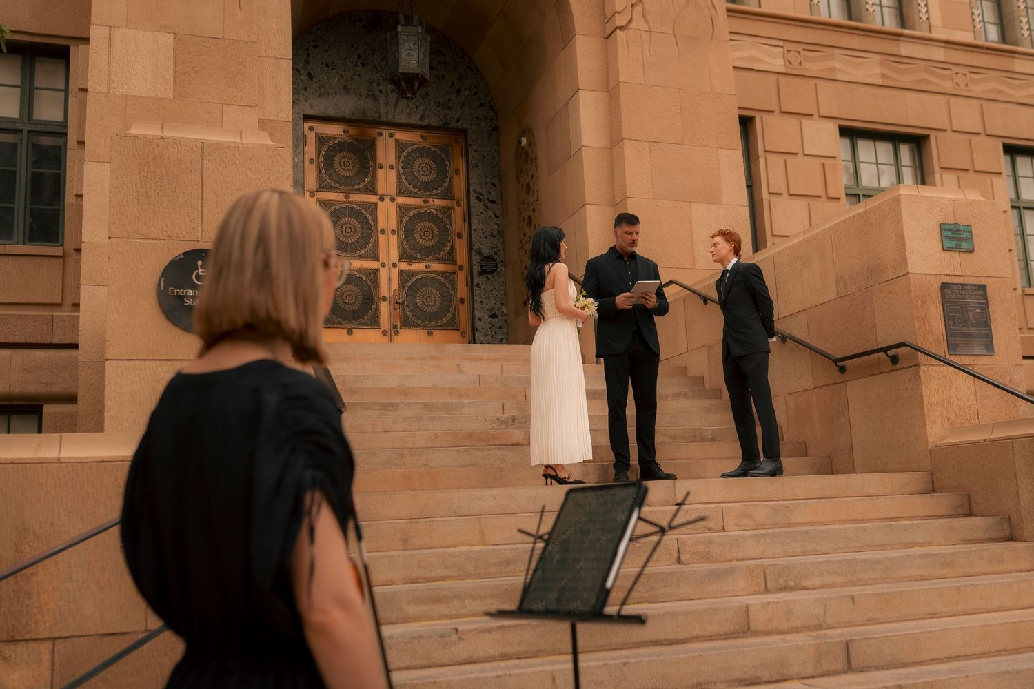 Couple exchanging vows on stone building steps with officiant and guest in black dress watching ceremony.