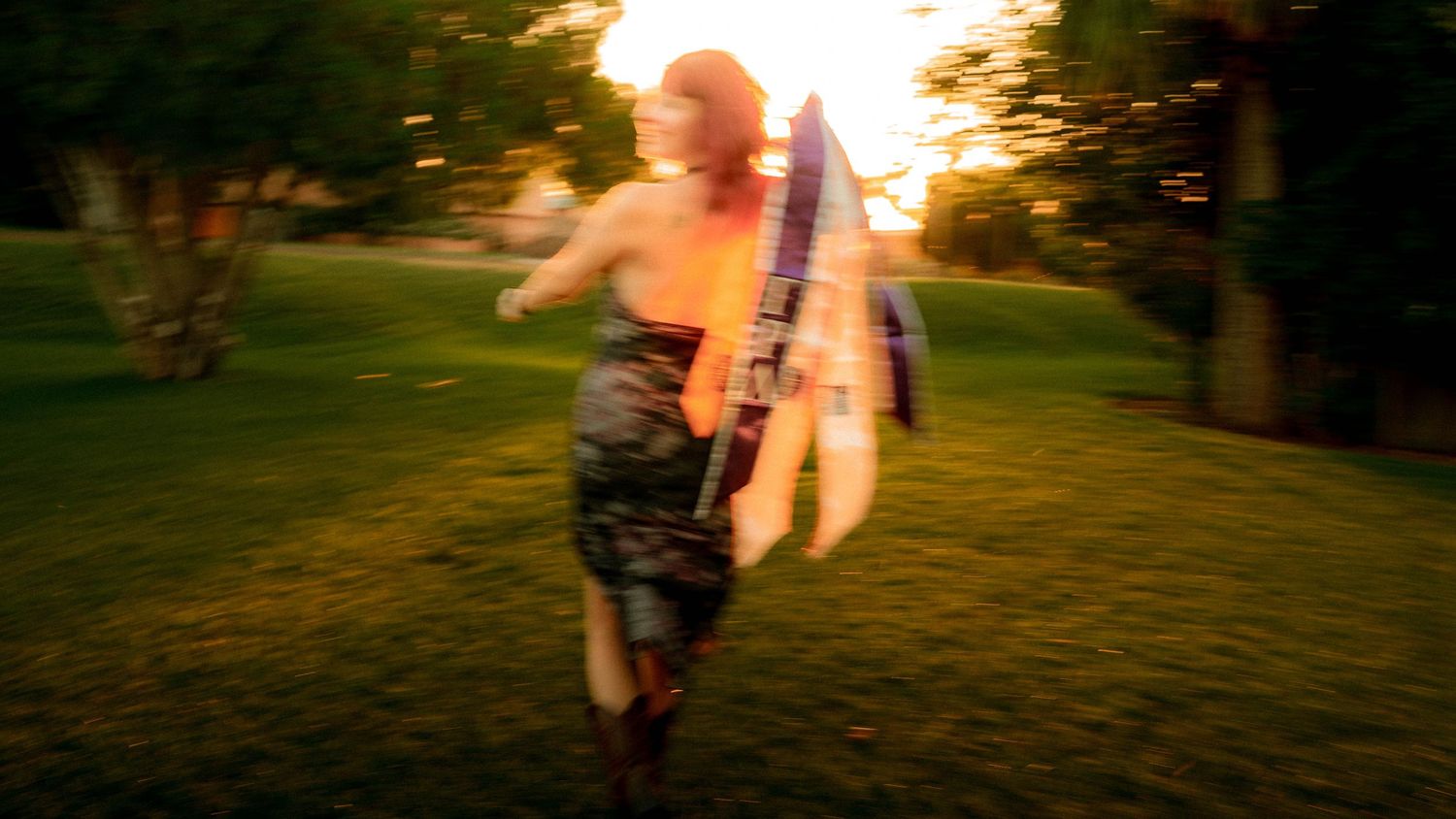 Person running across a green lawn at sunset, carrying a large guitar in warm golden light.