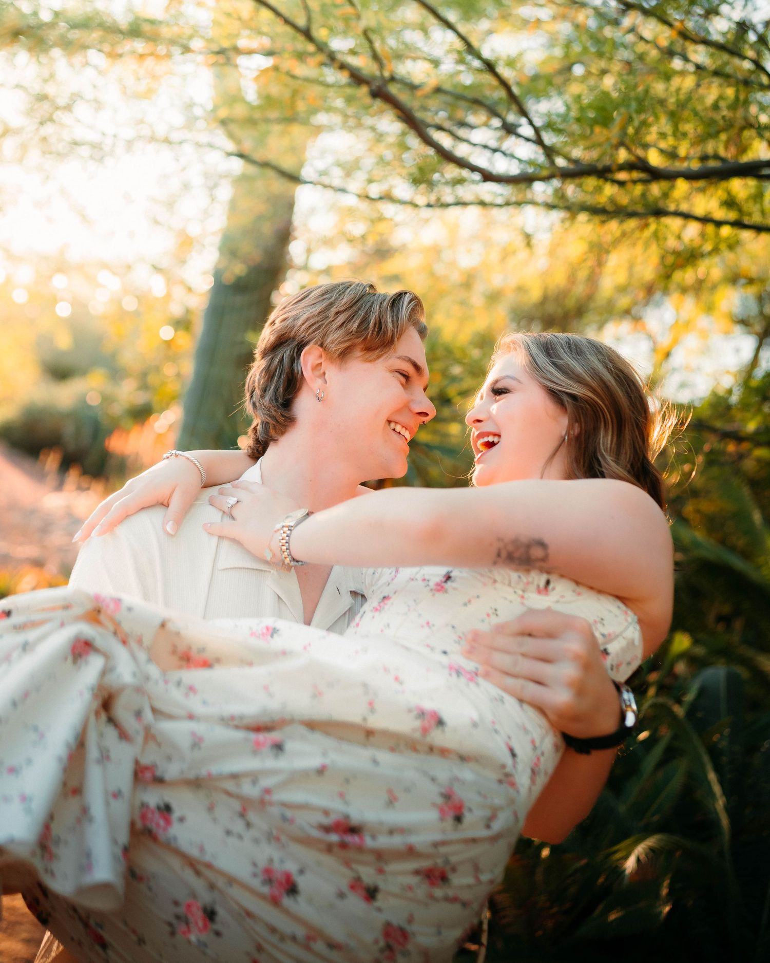 A couple shares a joyful moment as a man carries a woman in a floral dress outdoors in golden sunlight.