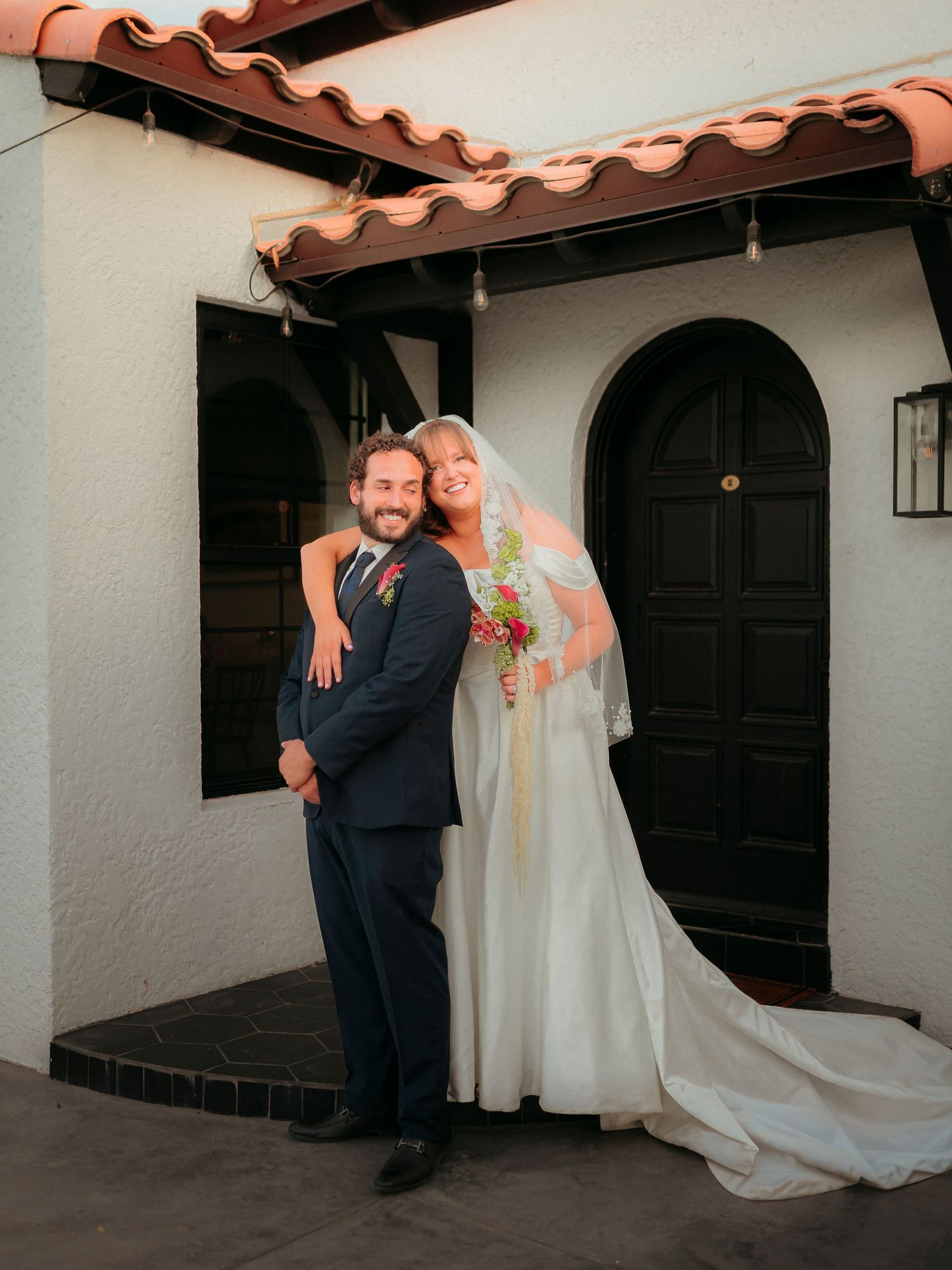 Bride and groom posing together in front of a white Spanish-style building with arched doors and terracotta roof tiles.