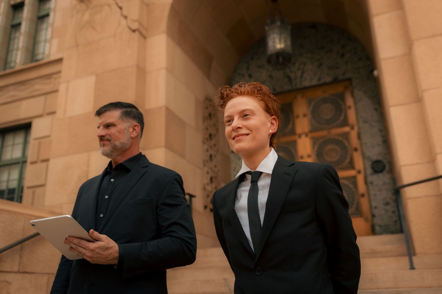 Young red-haired groom in black suit standing outside a stone building with officiant.